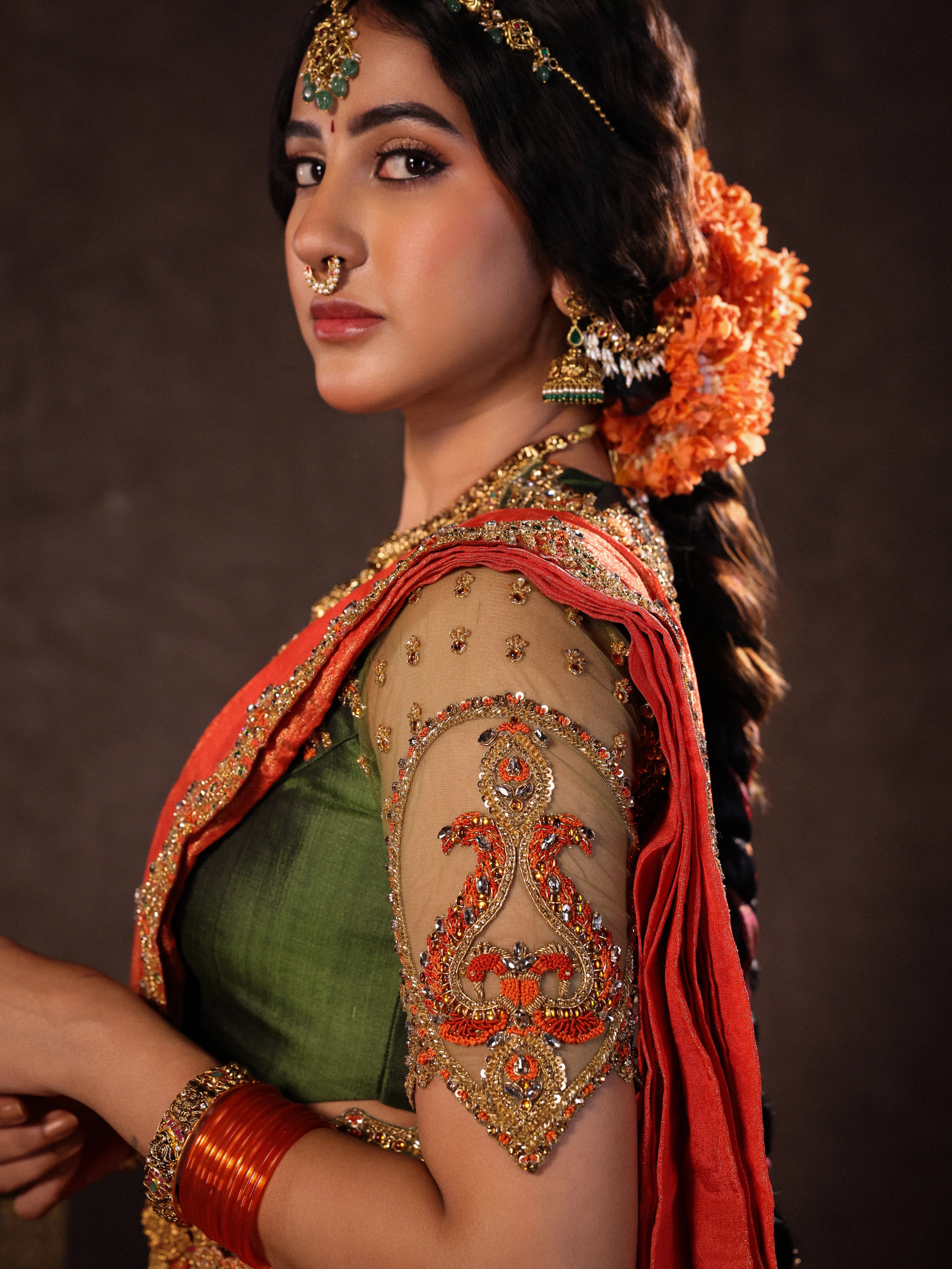 Actress Akshaya Hariharan during photoshoot in an orange & olive green silk half saree set with tulle sleeves. Handcrafted with zardozi, resham, cutdana, sequins & rhinestones. Team Archana Karthick completed the style with orange glass bangles, a ring, jhumkas, nose ring and a maang tikka.