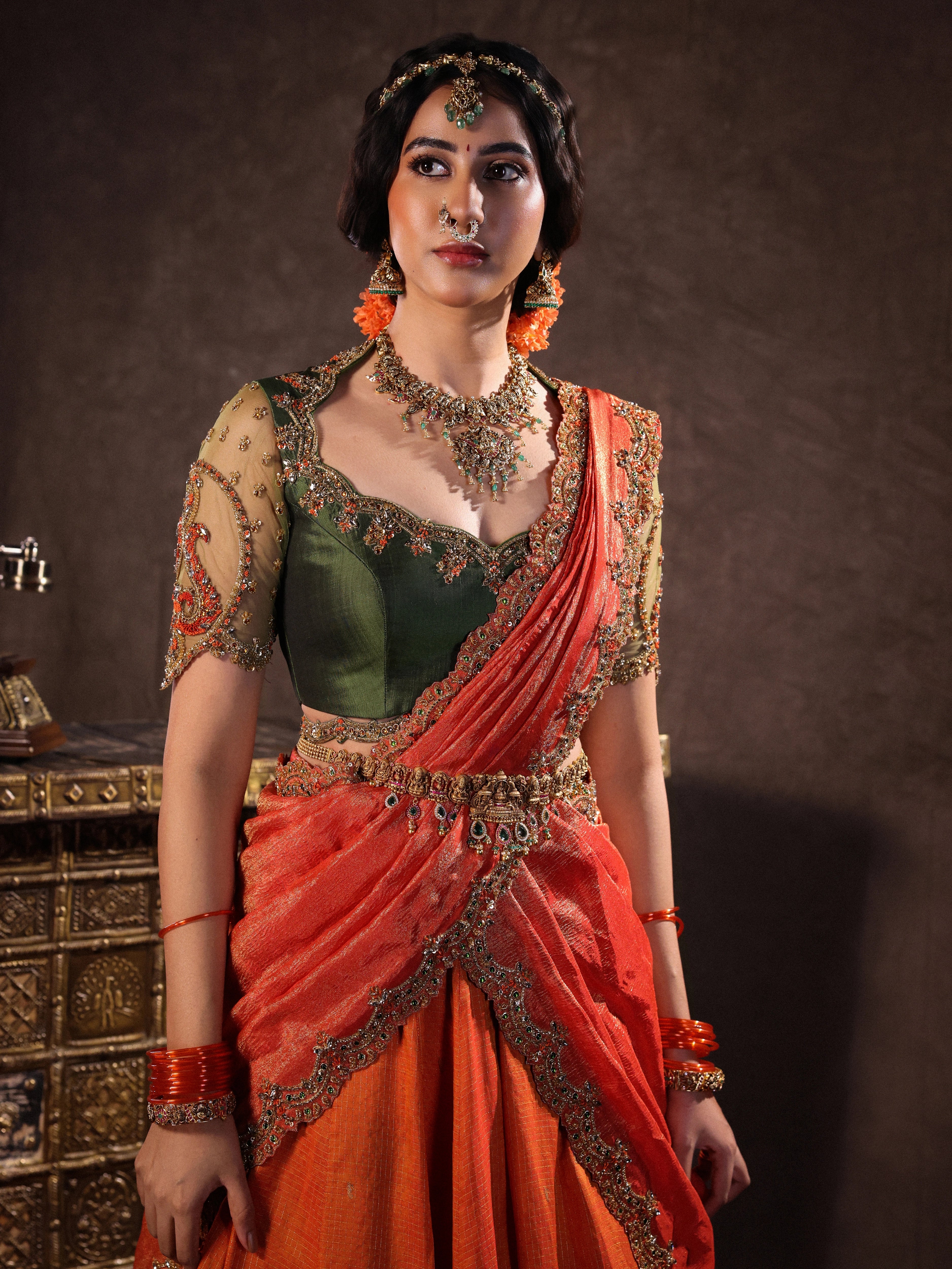 Actress Akshaya Hariharan for photoshoot in an orange & olive green silk half saree set with tulle sleeves. Handcrafted with zardozi, resham, cutdana, sequins & rhinestones. Team Archana Karthick completed the style with orange glass bangles, a ring, jhumkas, nose ring and a maang tikka.
