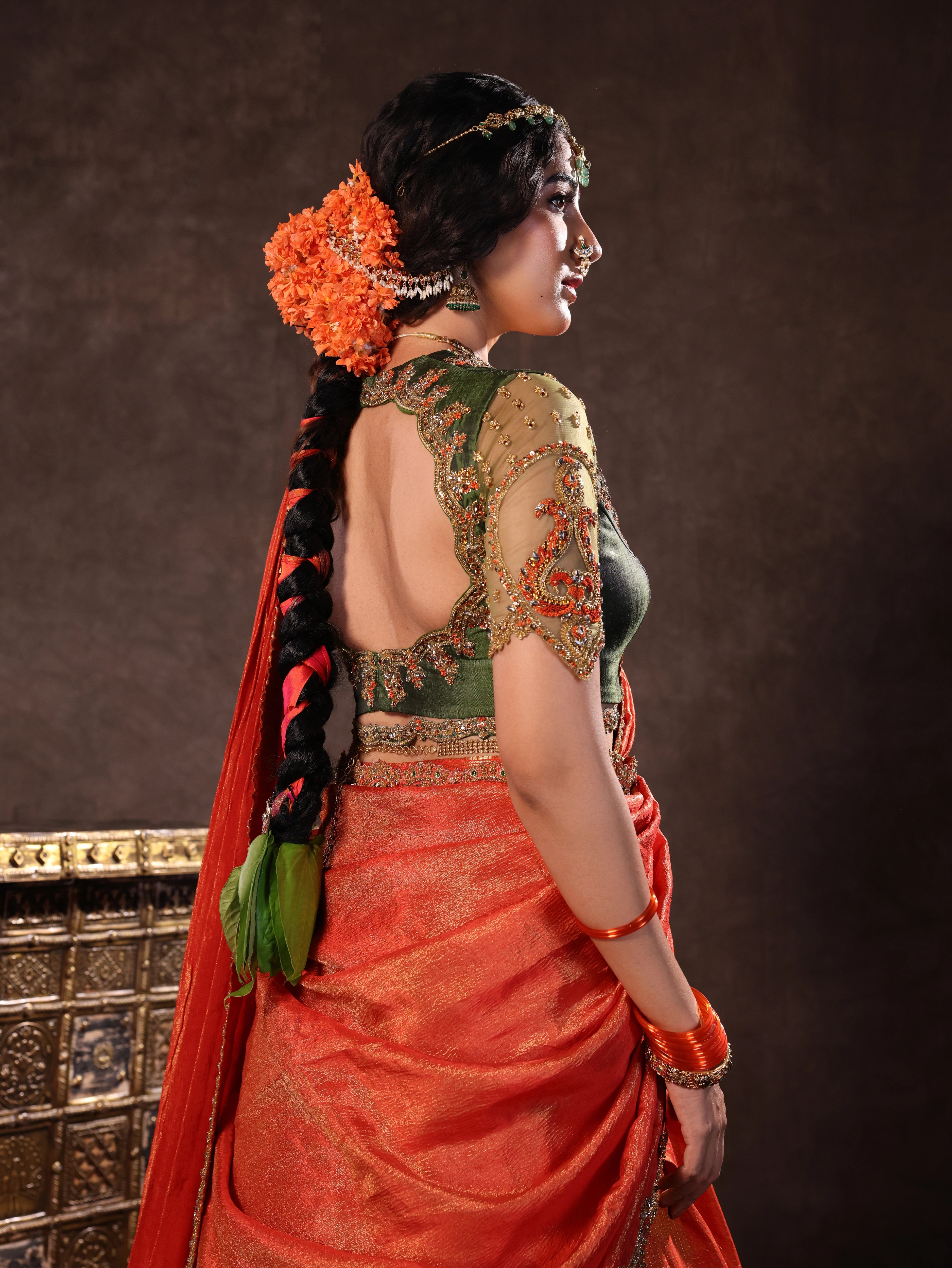 Actress Akshaya Hariharan poses for back design in an orange & olive green silk half saree set with tulle sleeves. Handcrafted with zardozi, resham, cutdana, sequins & rhinestones. Team Archana Karthick completed the style with orange glass bangles, a ring, jhumkas, nose ring and a maang tikka.