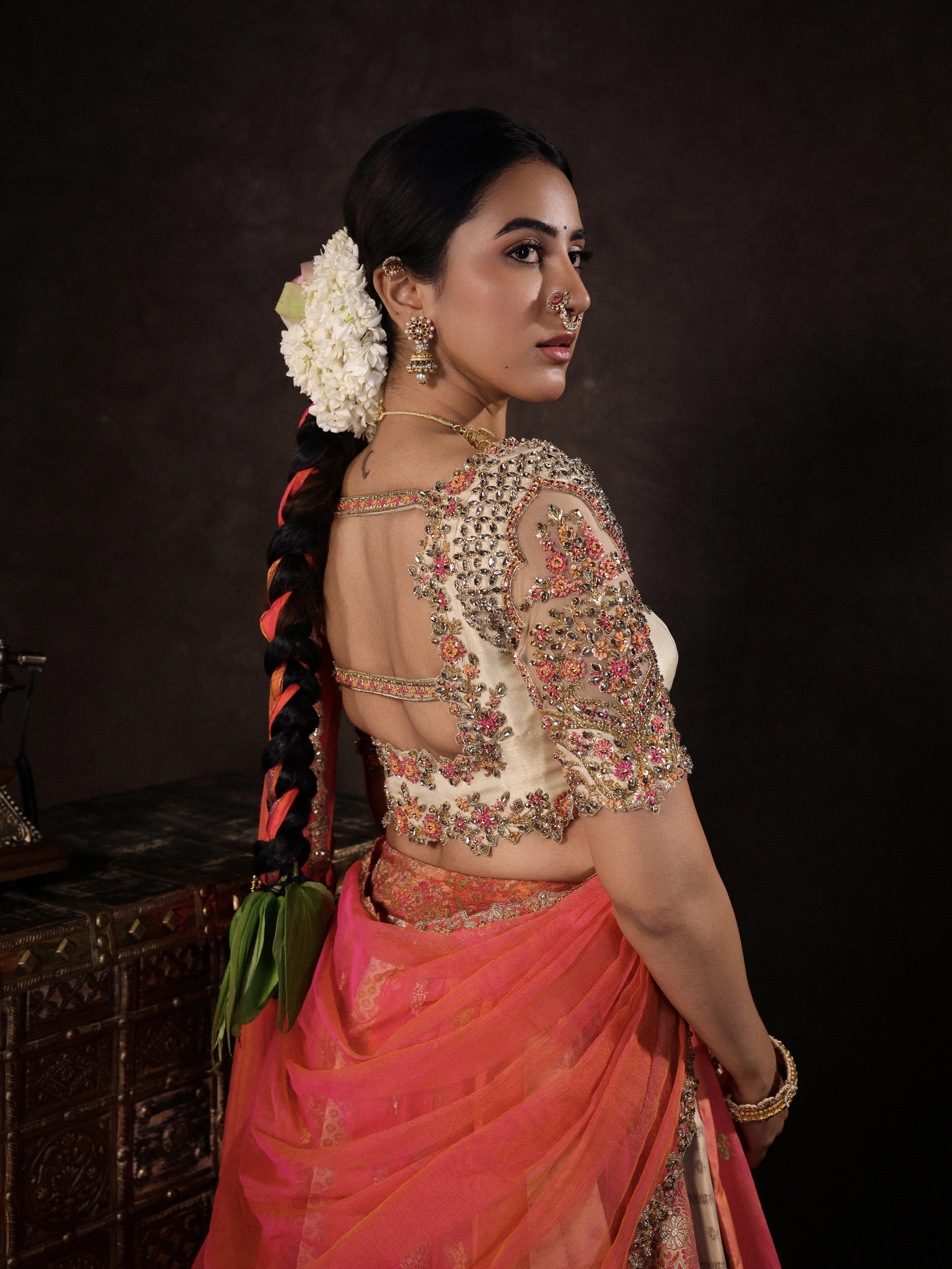 Actress Akshaya Hariharan posing for back blouse design in an ivory & pink brocade half saree set with tulle sleeves & dupatta. Team Archana Karthick completed the style with purple glass bangles, a ring, jhumkas, nose ring and a maang tikka.
