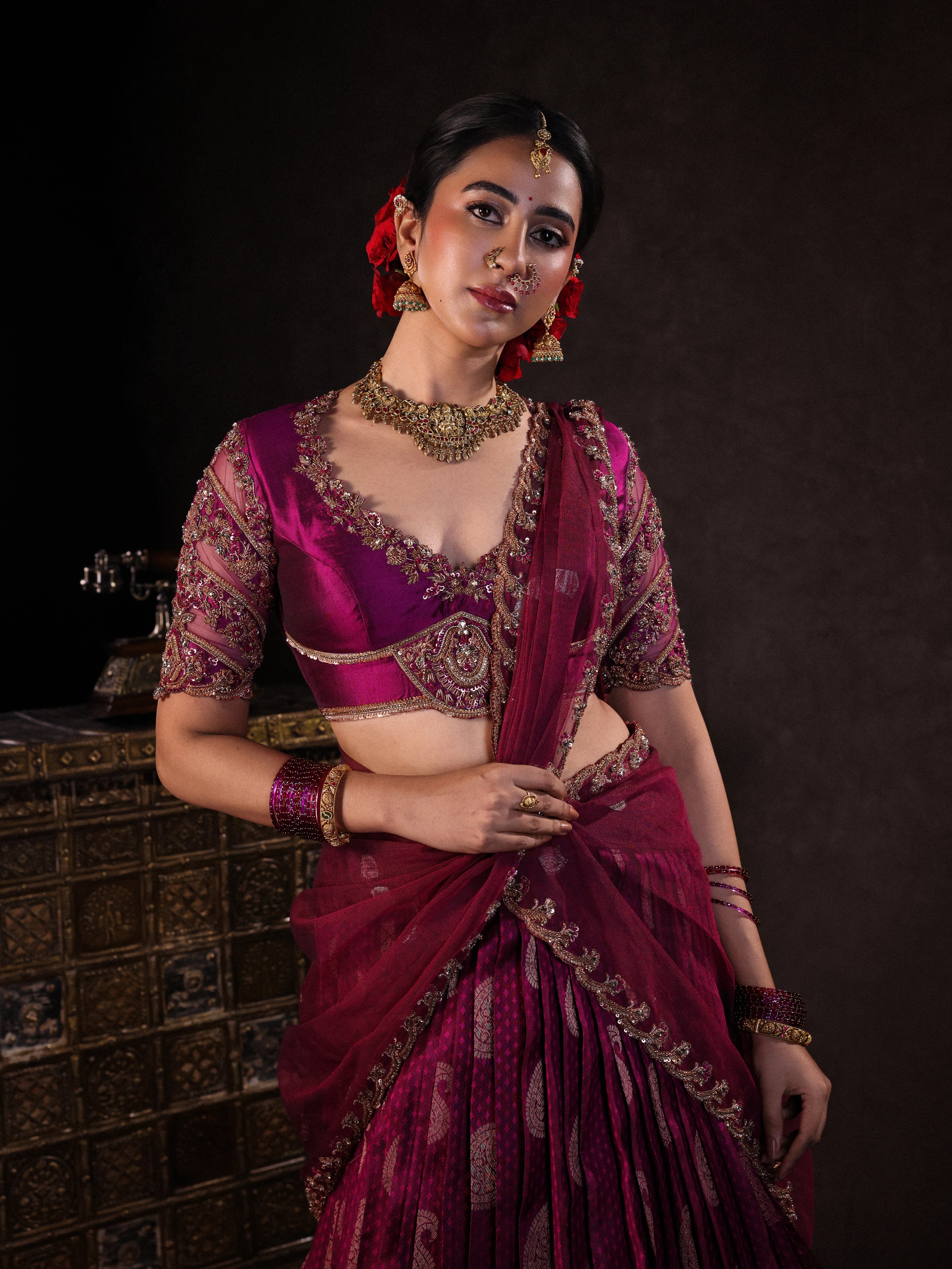 Actress Akshaya Hariharan posing for photoshoot in a dark magenta silk half saree set handcrafted with zardozi, resham, cutdana, sequins & rhinestones. Team Archana Karthick completed the style with dark magenta glass bangles, a ring, jhumkas, nose ring and a maang tikka.