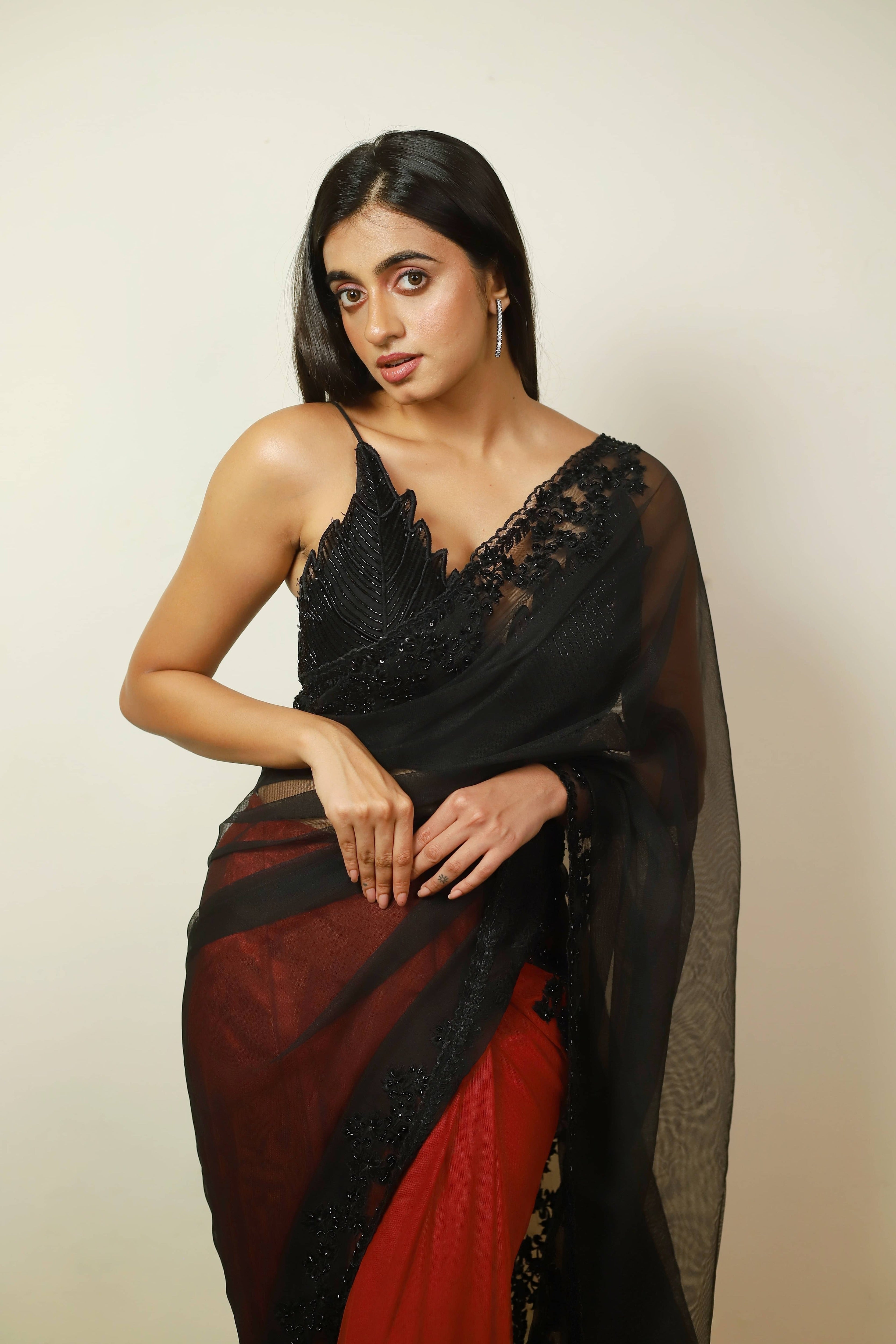 Actress Cherukuri Maanasa Choudhary who acted in the movie Lucky Baskhar with Dulquer Salmaan is seen in a red and black net saree with pallu and border detailings paired with a cut worked bralette with a plunge neck. Full front blouse profile.