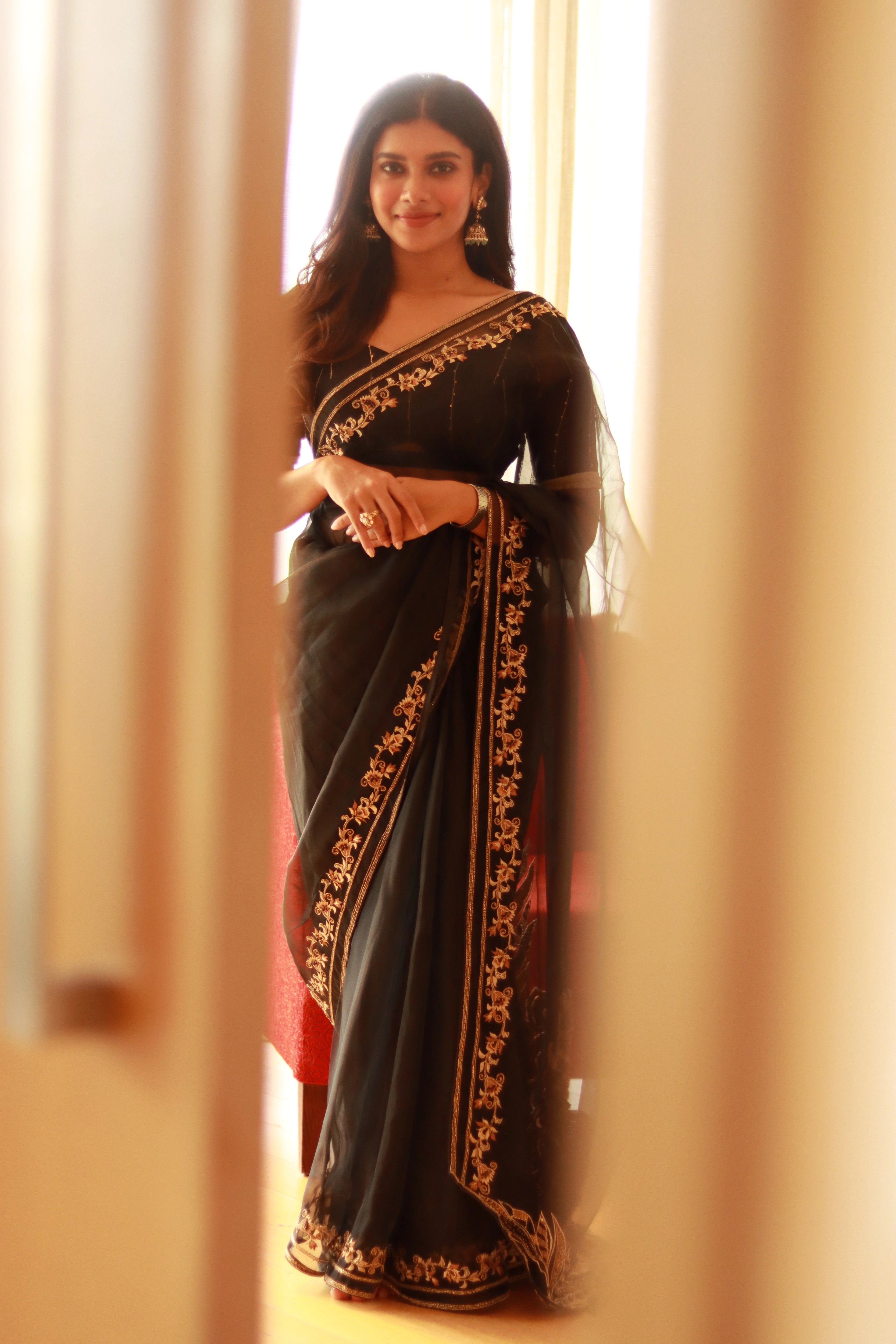 Actress Dushara Vijayan posing standing in a black organza saree, intricately handcrafted with cutdana, zari, beads, sequins and resham by team Archana Karthick.