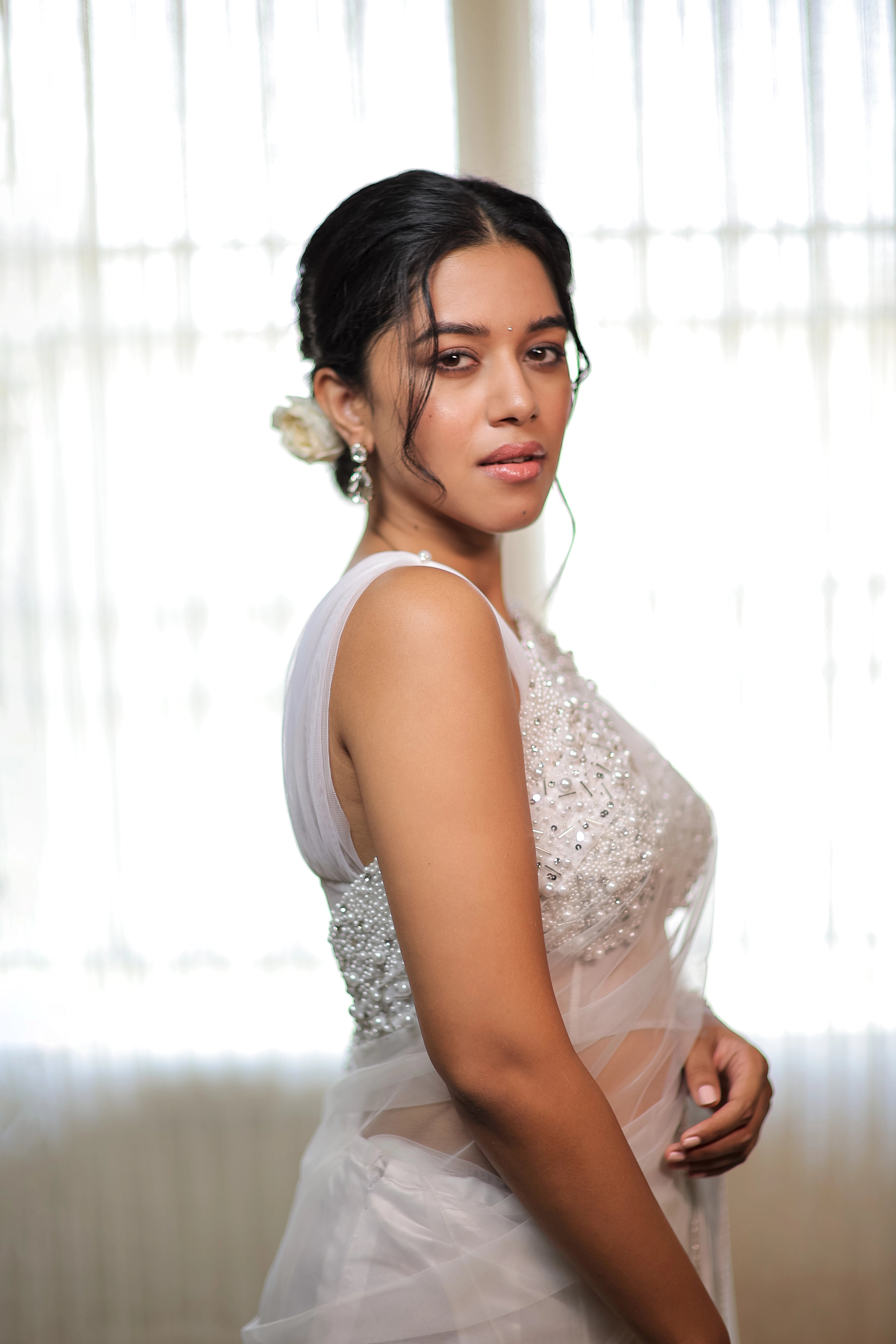 Actress Mirnalini Ravi posing for side profile in a pearl white saree intricately embellished with pearls and cut beads, paired with an off-shoulder corset with cowl sleeves.
