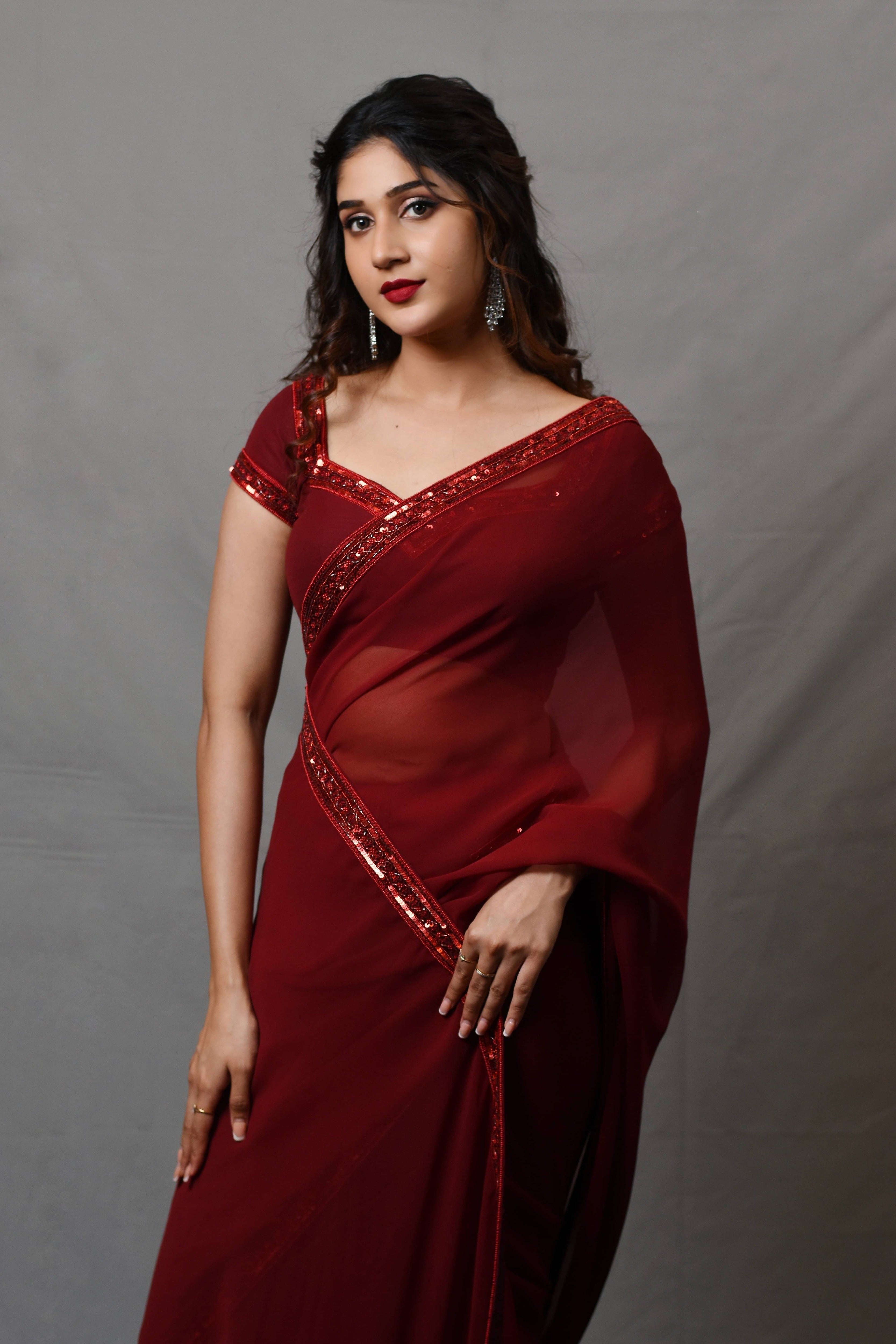 Actress Rabiya Khatoon in a ruby red georgette saree set designed by Archana Karthick. front profile with hand on thigh.