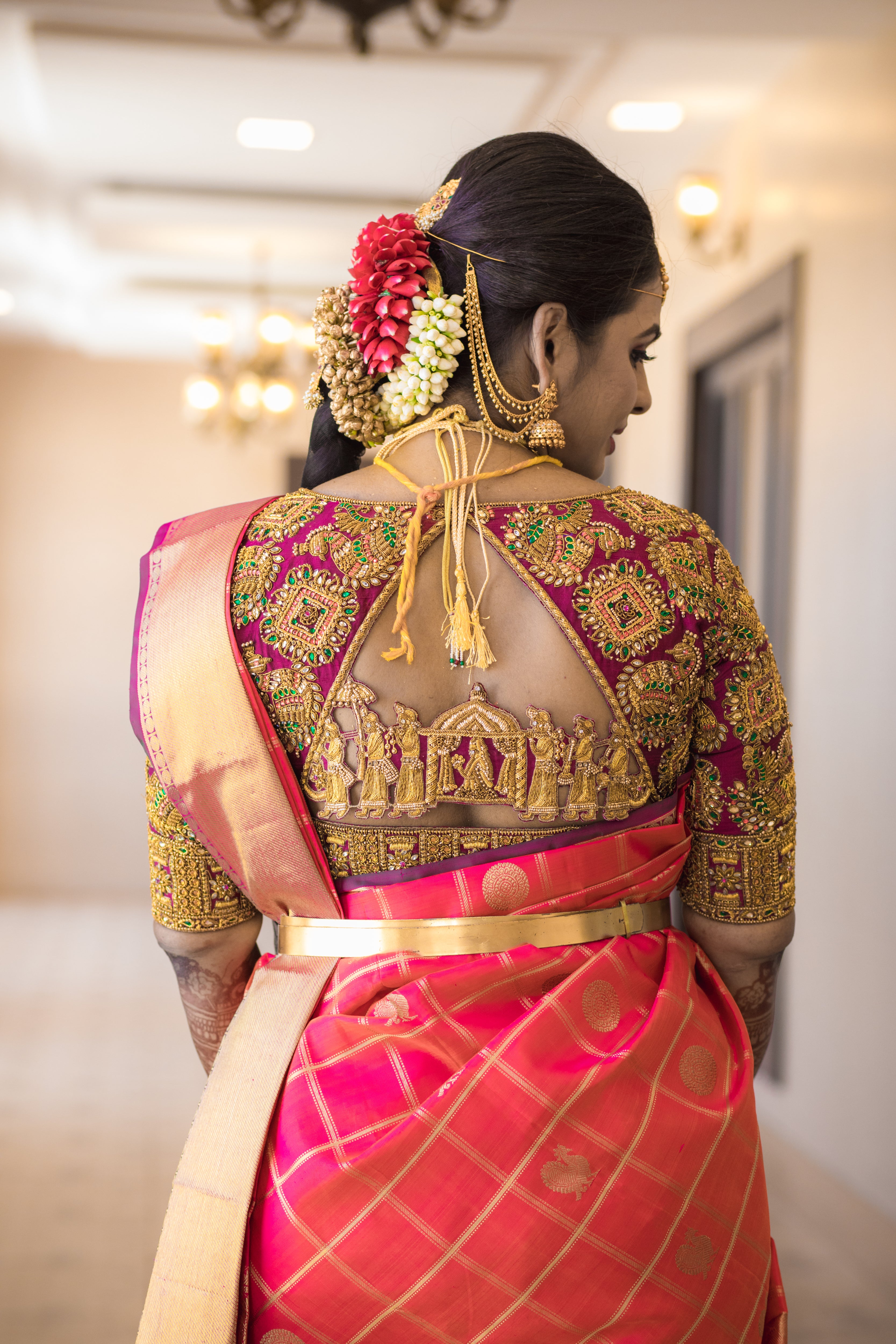 Archana Karthick Bride Kowsalya in a concept bridal blouse in purple raw silk. The blouse is heavily embellished with zardozi, kemp stones, beads and is half or elbow sleeved. The bride is posing in a way to show off the palanquin work at the back of the blouse which is a materpiece of art.