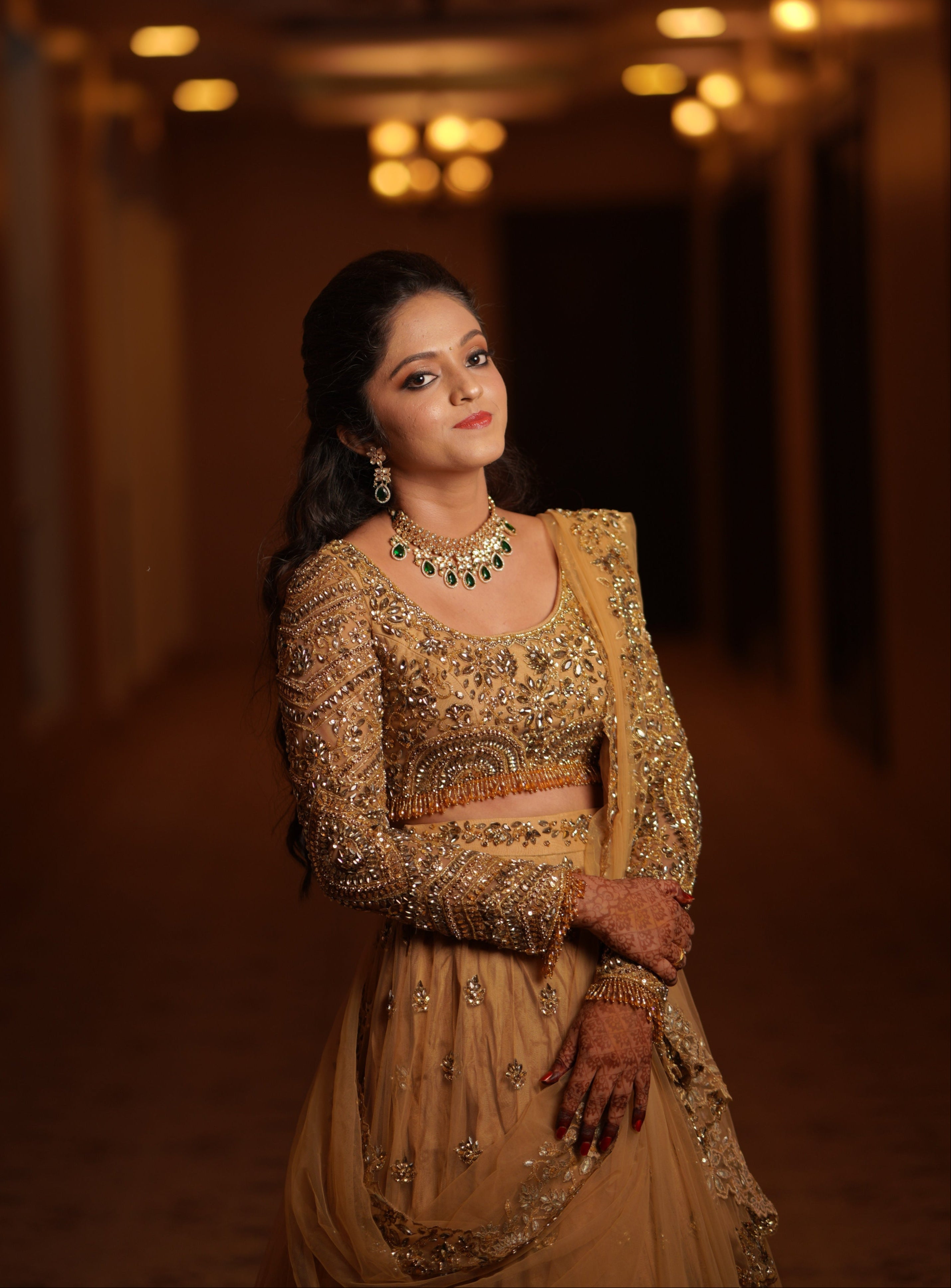 Archana Karthick Bride in golden beige lehenga with stonework poses for a front and side half profile. The intricate work on the full sleeved blouse inspired by mandala motifs is clearly breathtaking. The outfit has cutworked borders or seams of the dupatta (veil) and of the skirt. The seams of the blouse are embellished with crystal drops or stones. The blouse look stunning with rhinestone work, crystals and cut dana work.