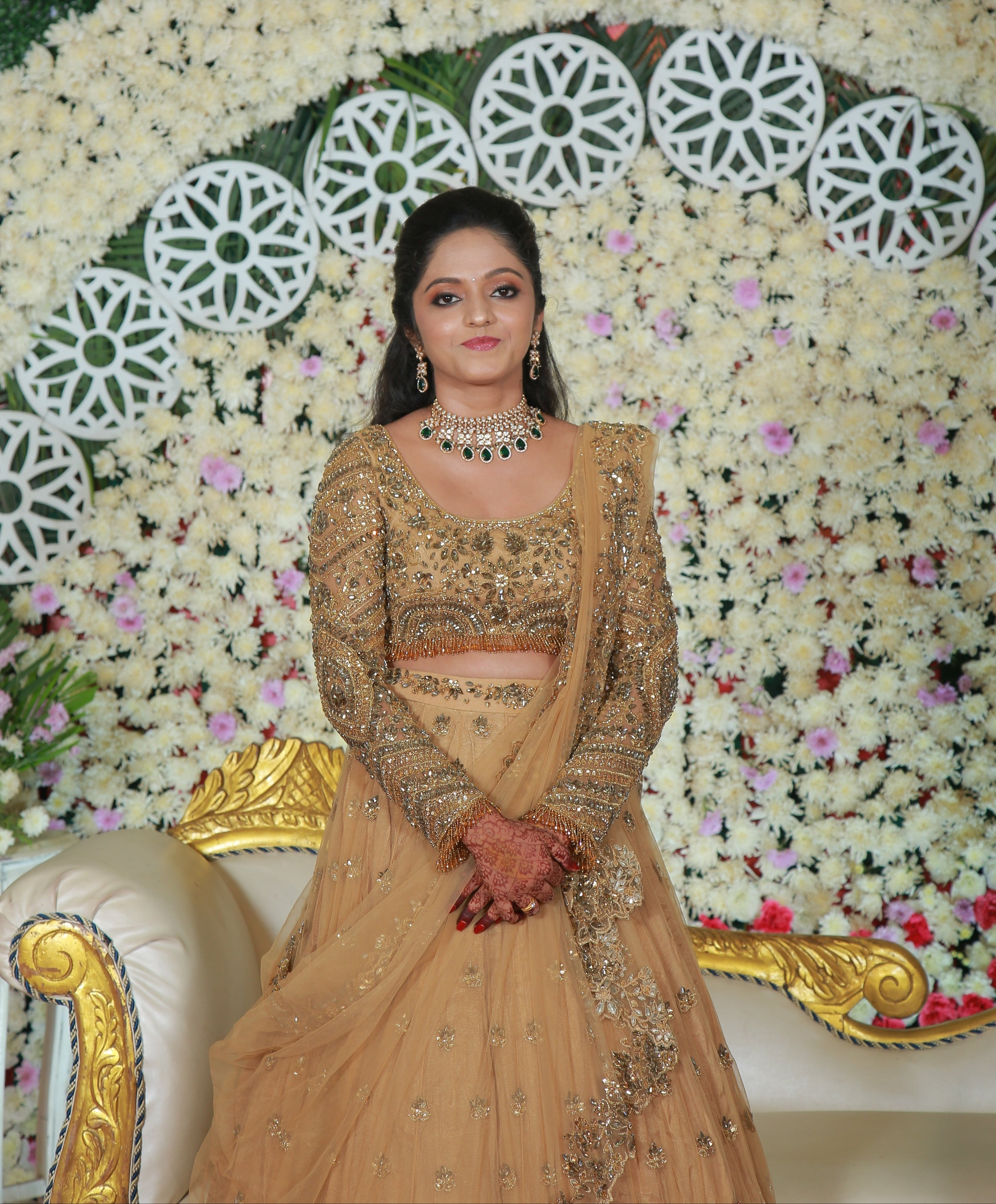 Archana Karthick Bride in golden beige lehenga with stonework posing for front profile on her reception stage which has a backdrop of white and lavender flowers. The outfit has cutworked borders on the seams of the dupatta (veil) and of the skirt. The seams of the blouse are embellished with crystal drops or stones. The mandala motifs on the blouse look stunning with rhinestone work, crystals and cut dana work.