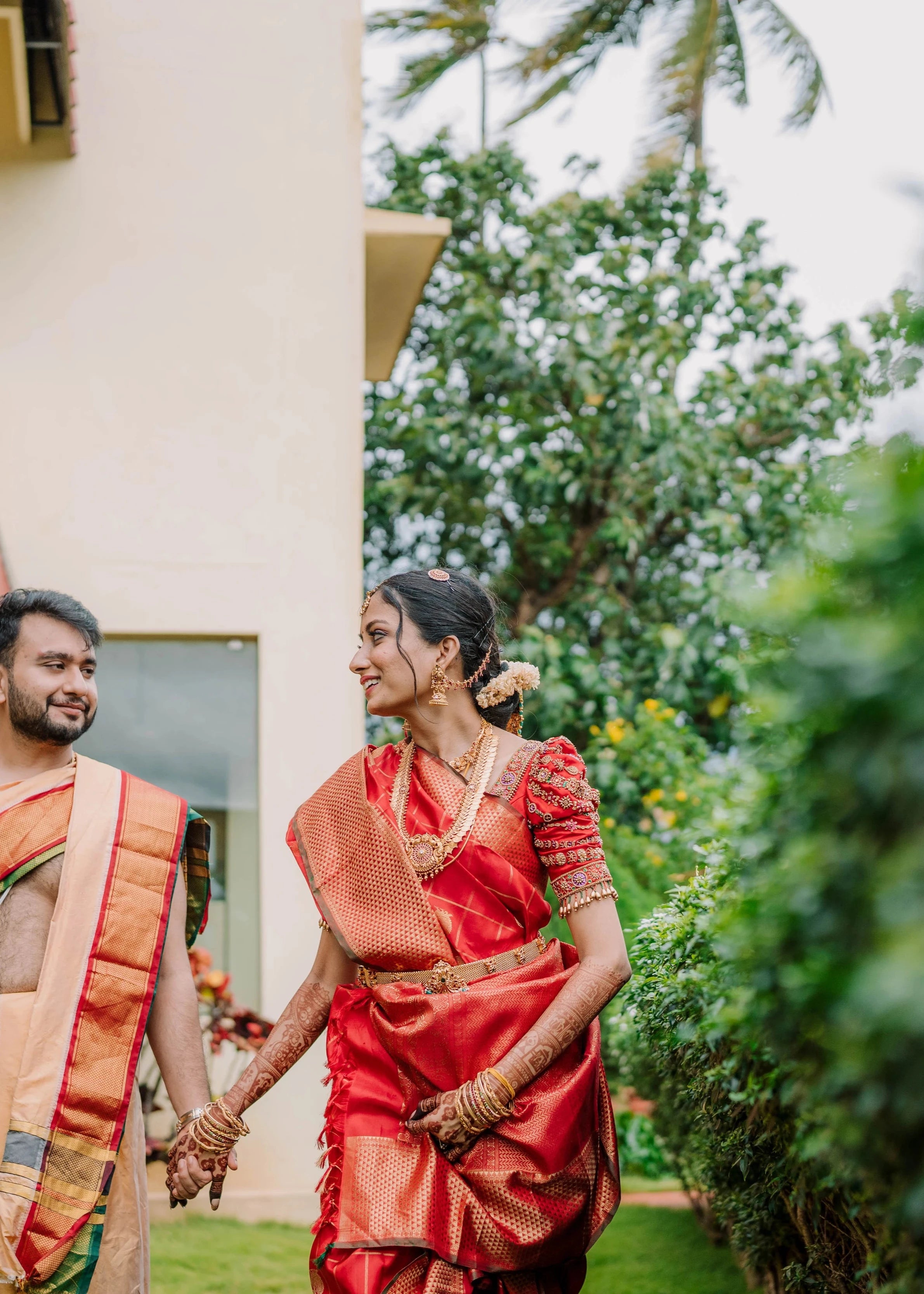 Bride Anusha K walking with groom in a red concept silk puff sleeved blouse, intricately handcrafted with zardosi, beads, kundan, and kemp stones by team Archana Karthick.