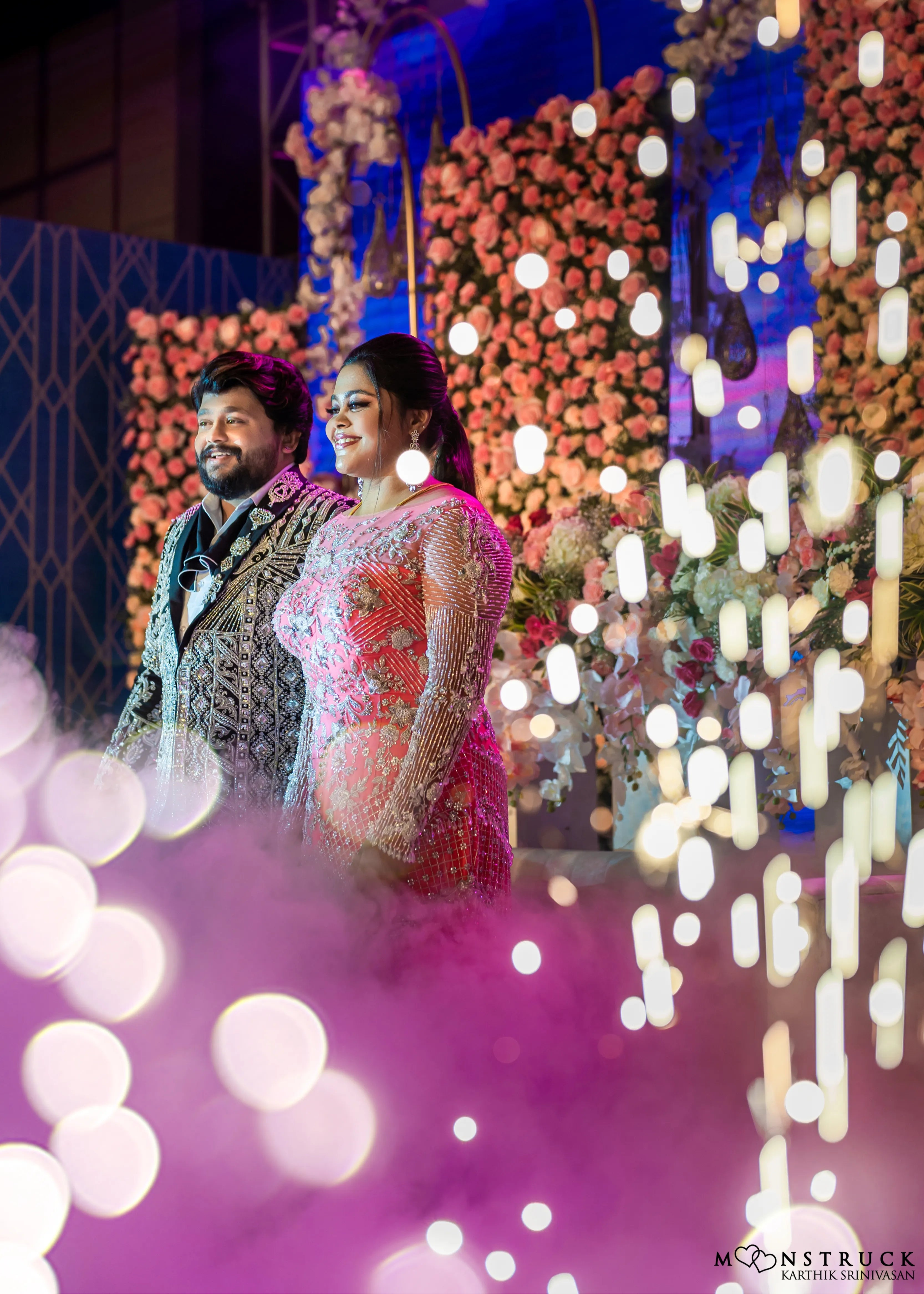 Bride (Coimbatore) in handcrafted pink tulle gown and and Groom (Chennai) in embeliished jacket for their reception smiling at the guests.