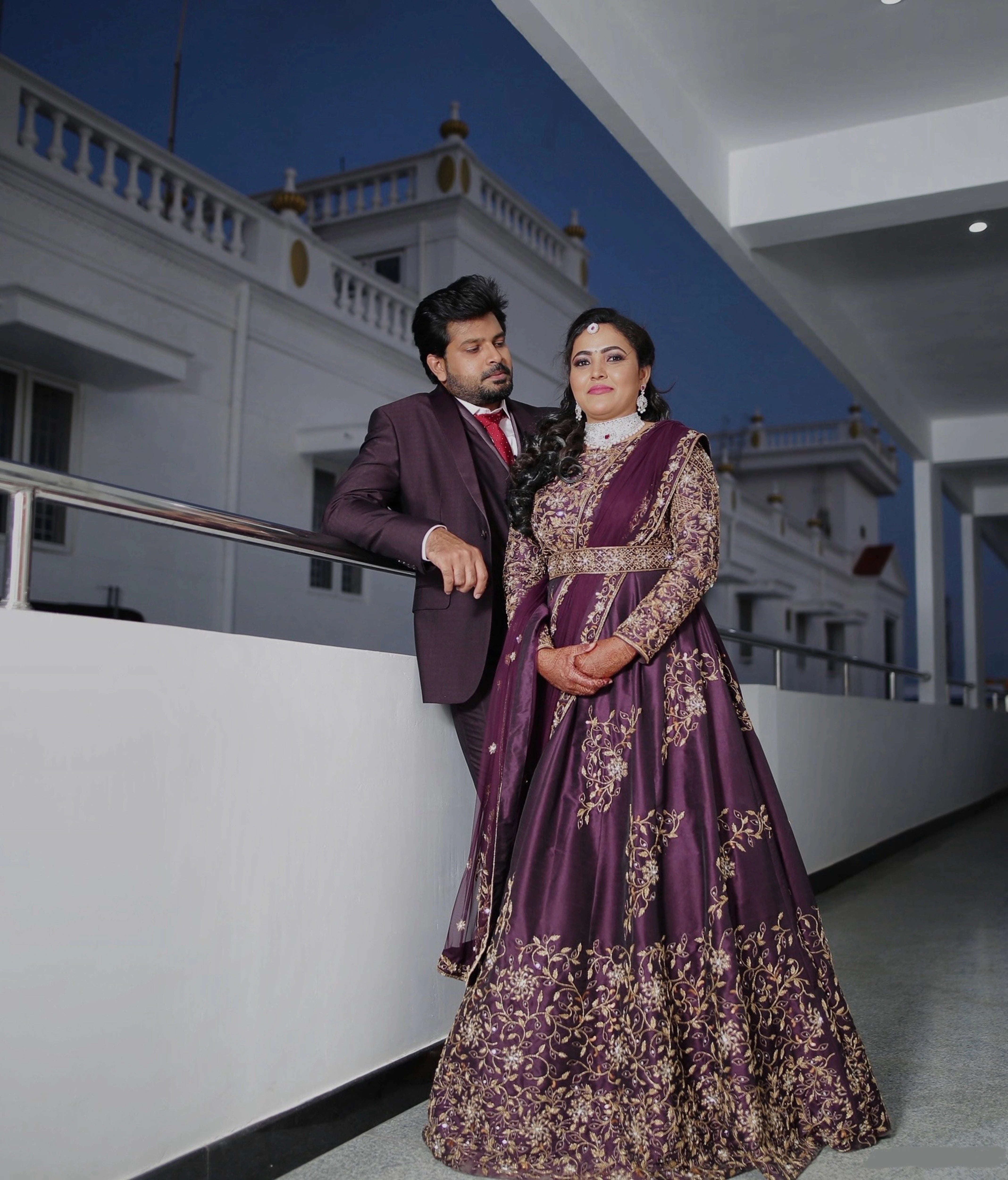 Bride Deepika posing with groom in an eggplant colored raw silk draped gown with belt intricately embellished with cutdana, sequins, stones and beads by team Archana Karthick.