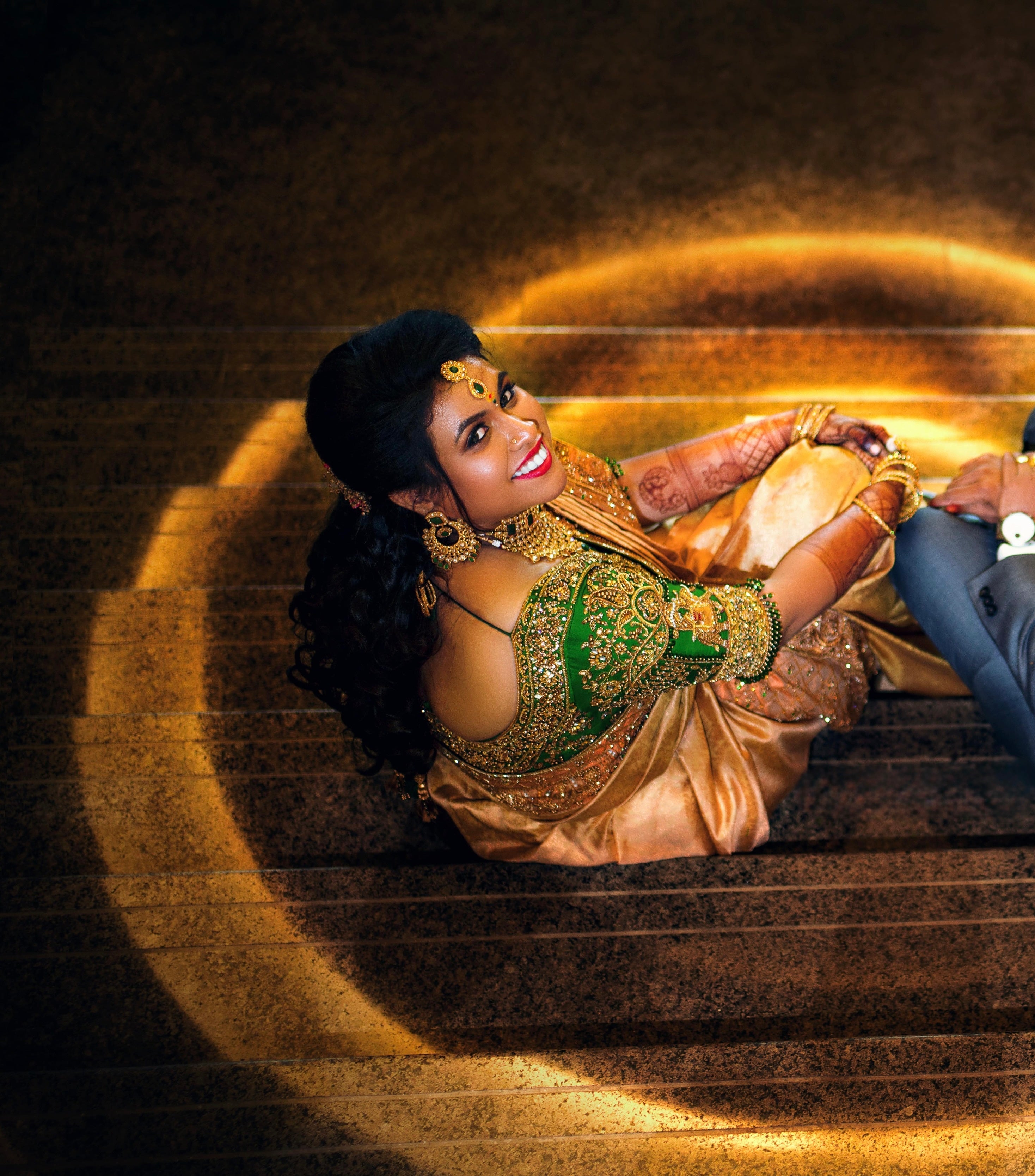 Bride Hemakiruthika from Chennai in a green raw silk blouse and gold silk saree with heavy border embellishments, designed by Archana Karthick. She is looking to her right and is posing for an aerial shot