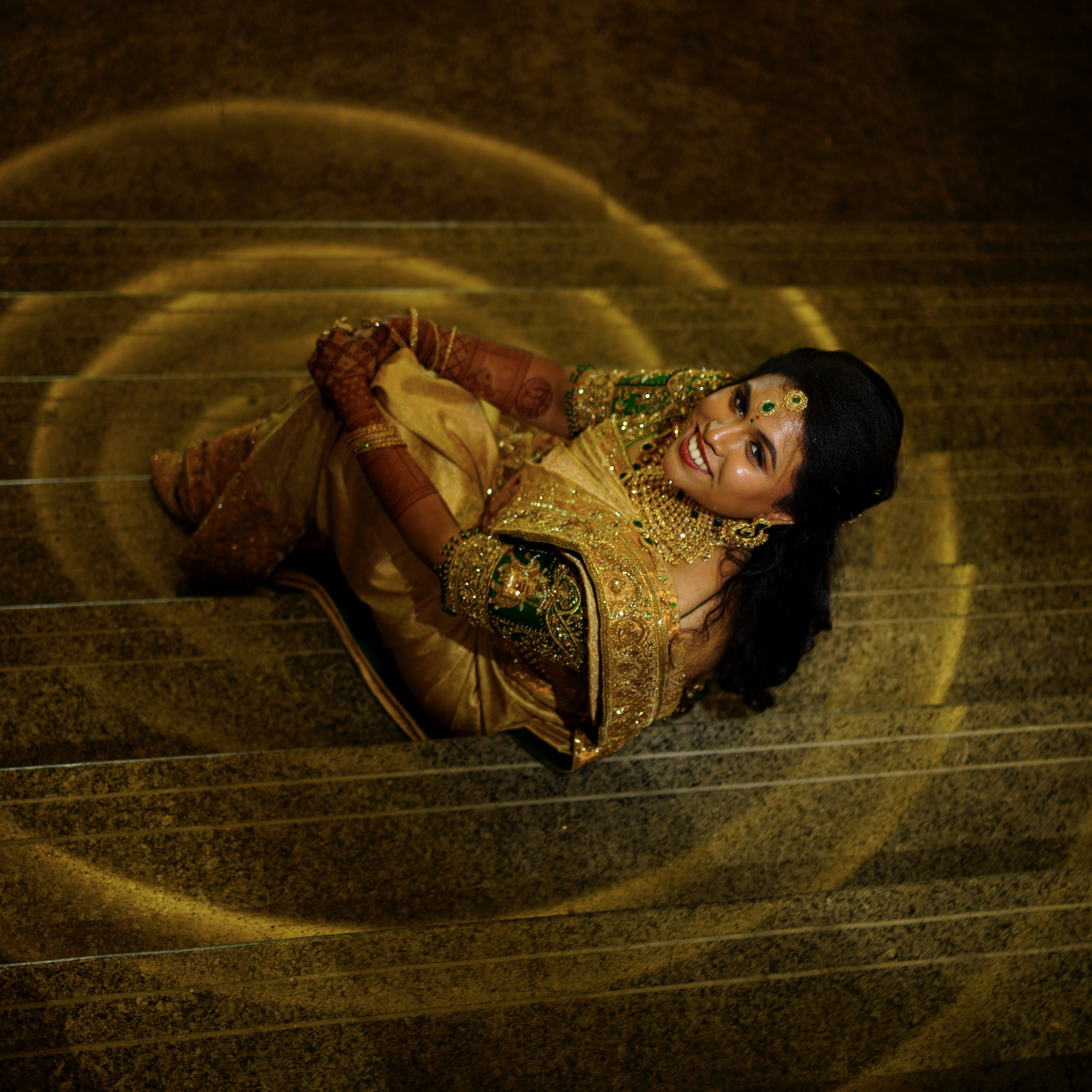 Bride Hemakiruthika from Chennai in a green raw silk blouse and gold silk saree with heavy border embellishments, designed by Archana Karthick. She is seated and is looking above as it is an aerial shot.