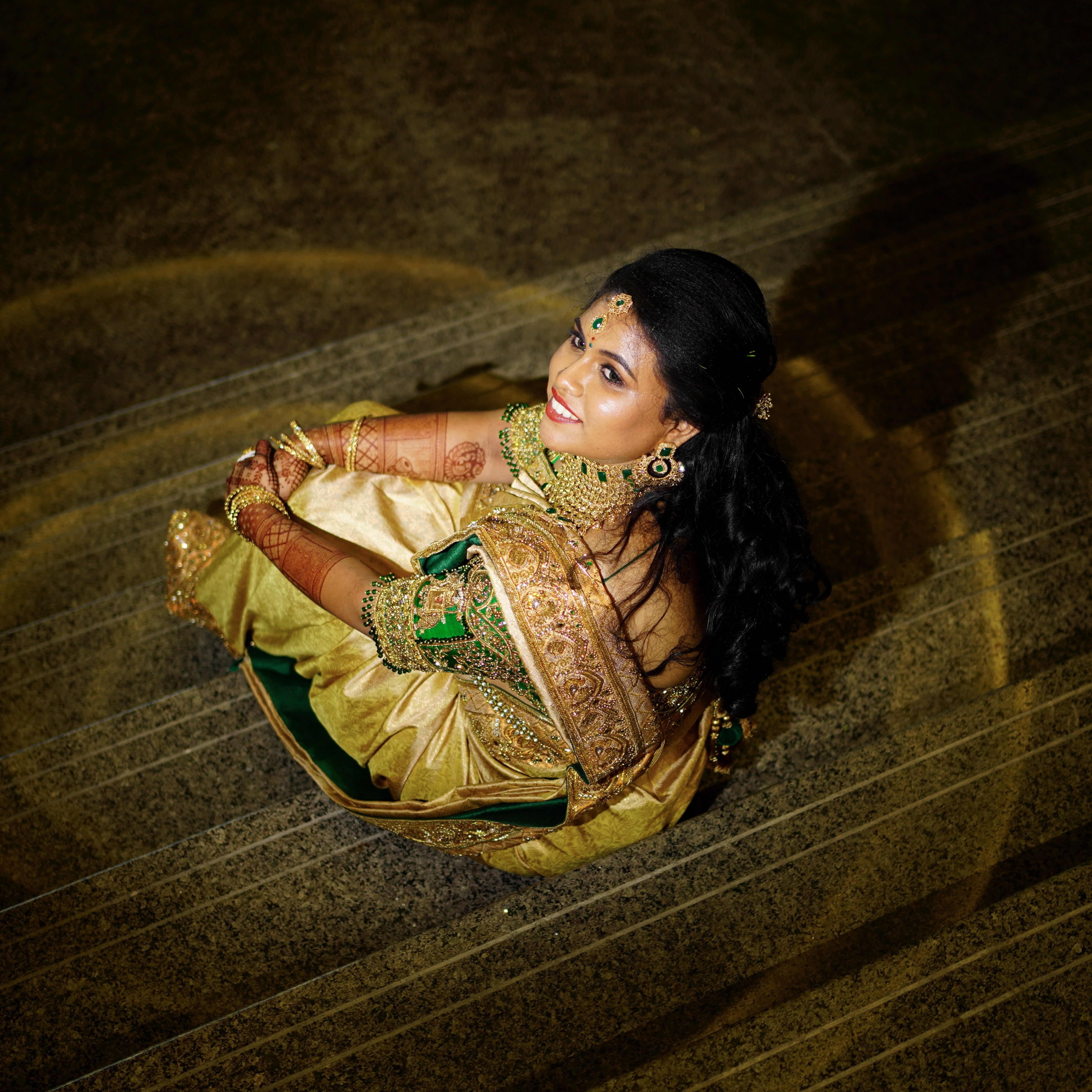 Bride Hemakiruthika from Chennai in a green raw silk blouse and gold silk saree with heavy border embellishments, designed by Archana Karthick. She is seated and is looking above as it is an aerial shot to her left.
