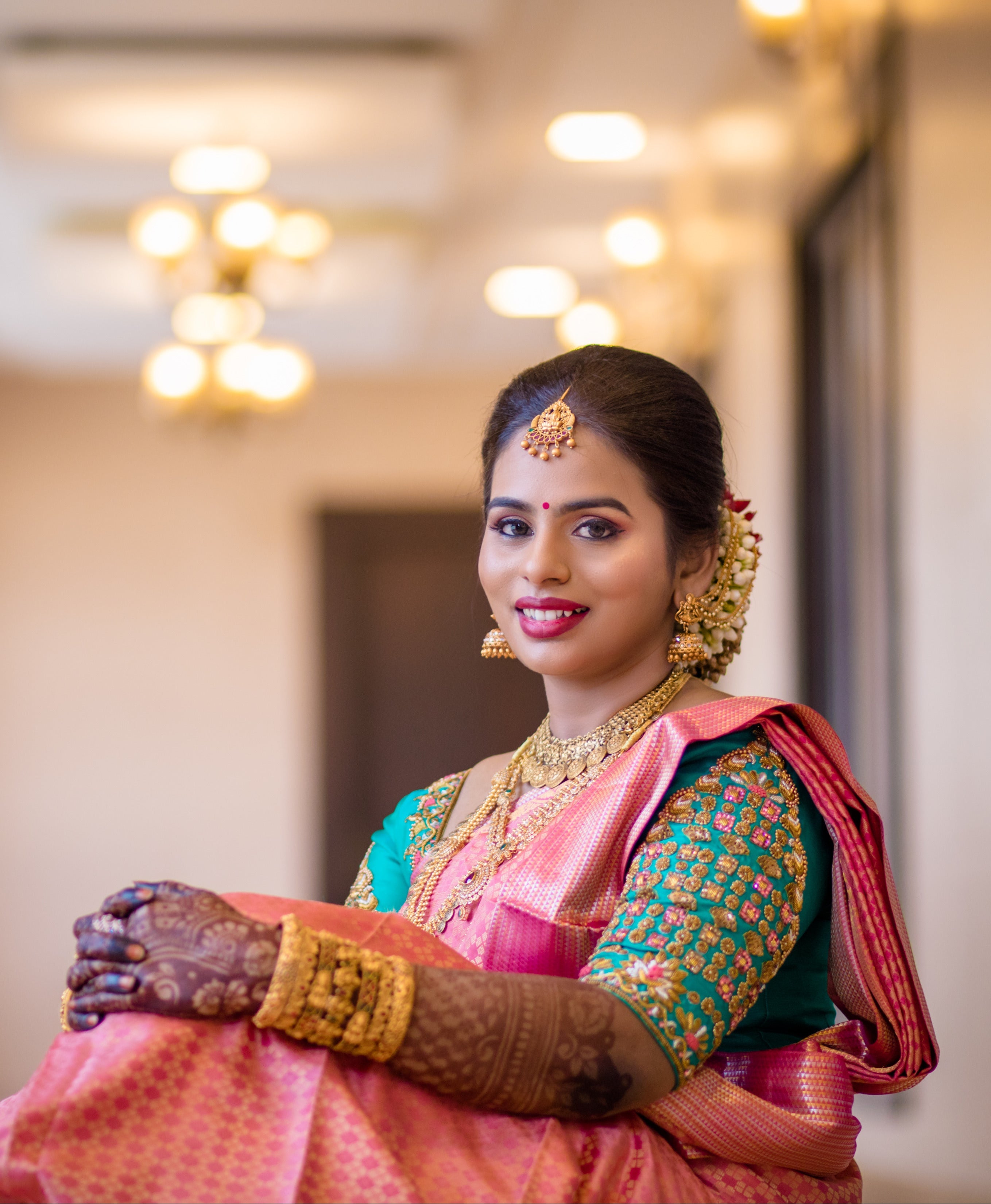 Bride Kowsalya from USA in a teal raw silk blouse designed by Archana Karthick. Posing seated and looking straight at the camera.