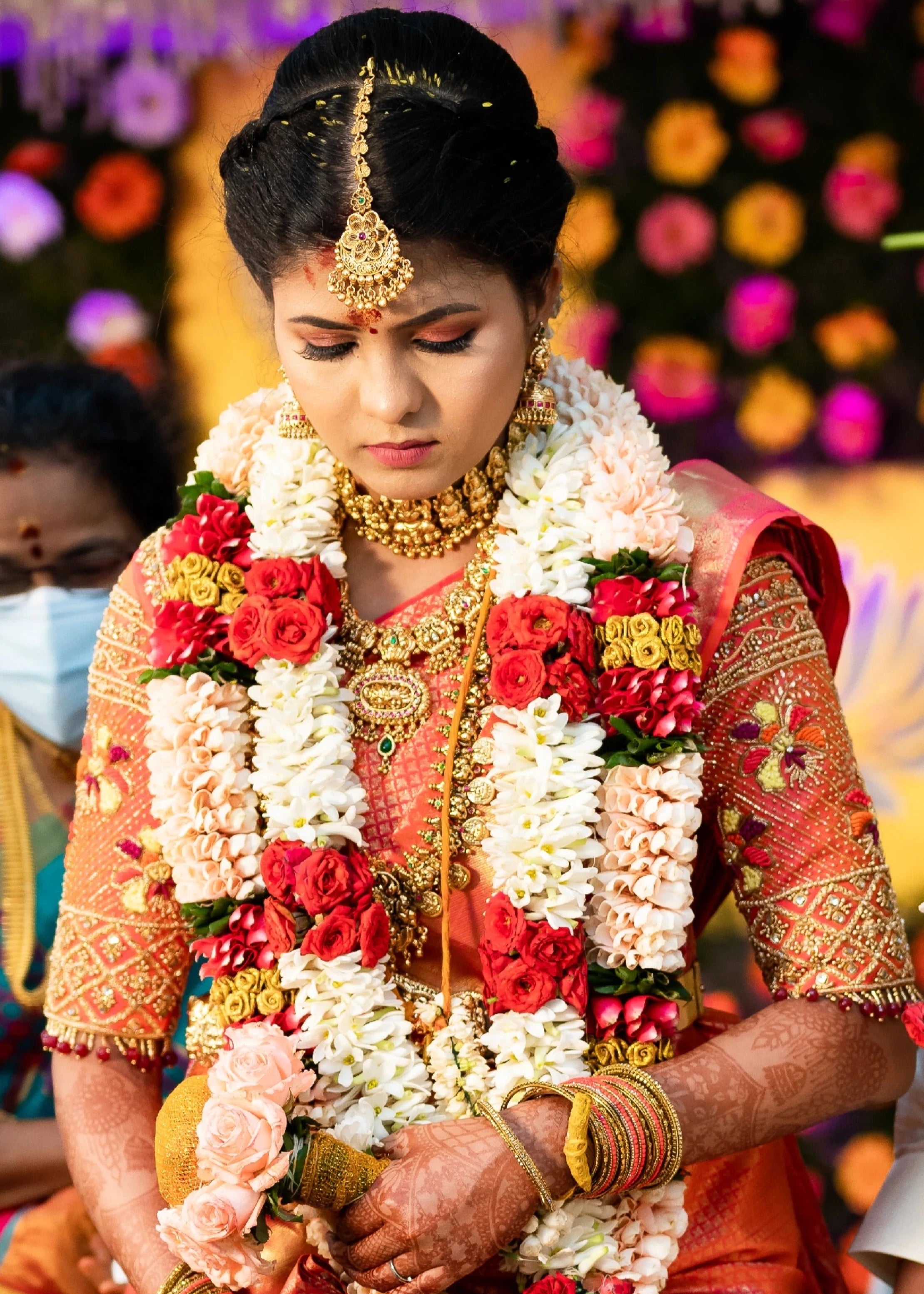 Bride Sarumathi looking down in an orange silk blouse, intricately handcrafted with zardozi, resham, stones and beads by team Archana Karthick.