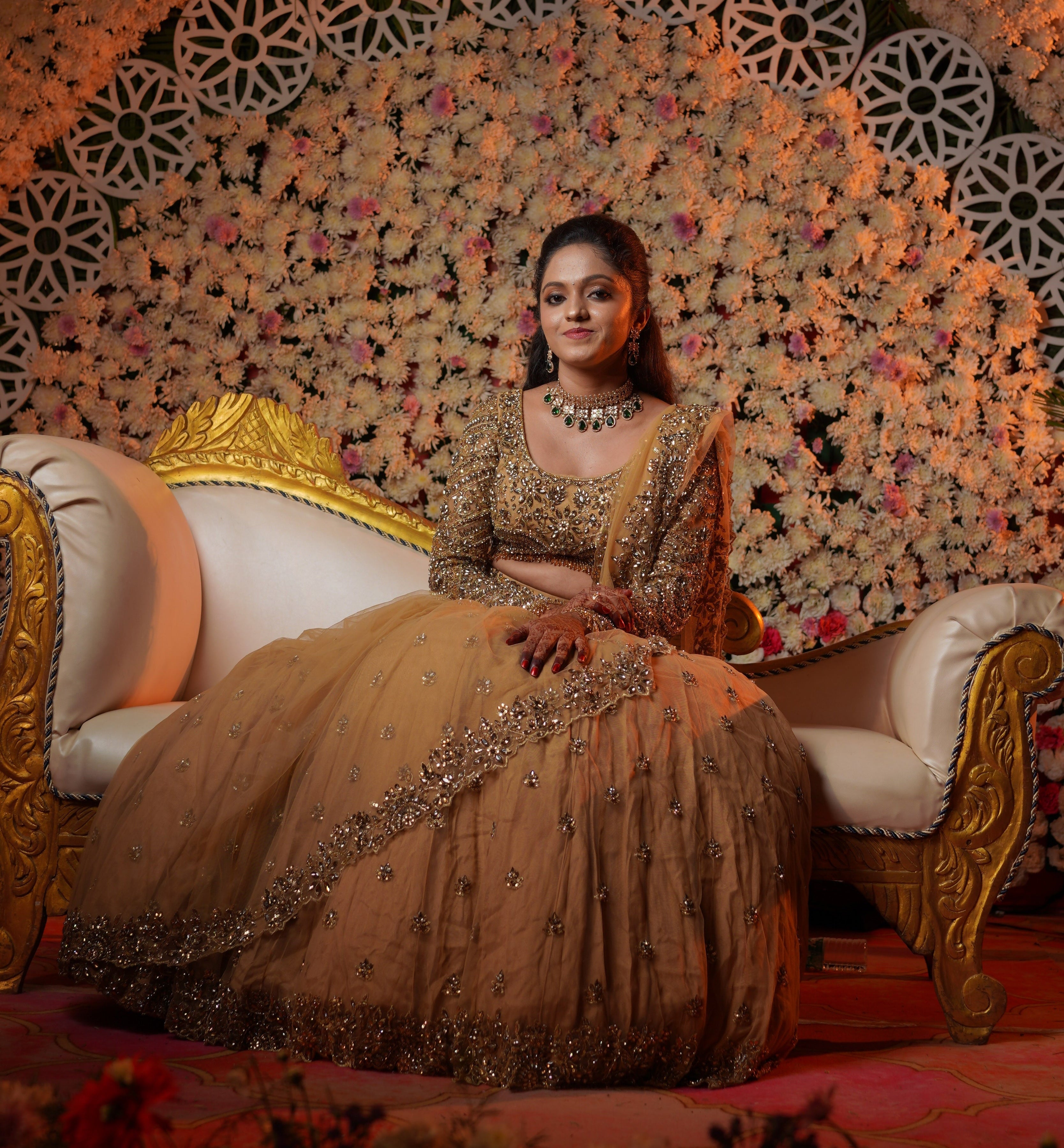 Bride Shravanthi in golden beige stonework lehenga is sitting on the couch on the stage during her recption. She smiles at the camera. Her Gold Lehnega in Net, looks very elegant. The outfit has cutworked borders or seams of the dupatta (veil) and of the skirt. The seams of the blouse are embellished with crystal drops or stones. The mandala motifs on the blouse look stunning with rhinestone work, crystals and cut dana work.
