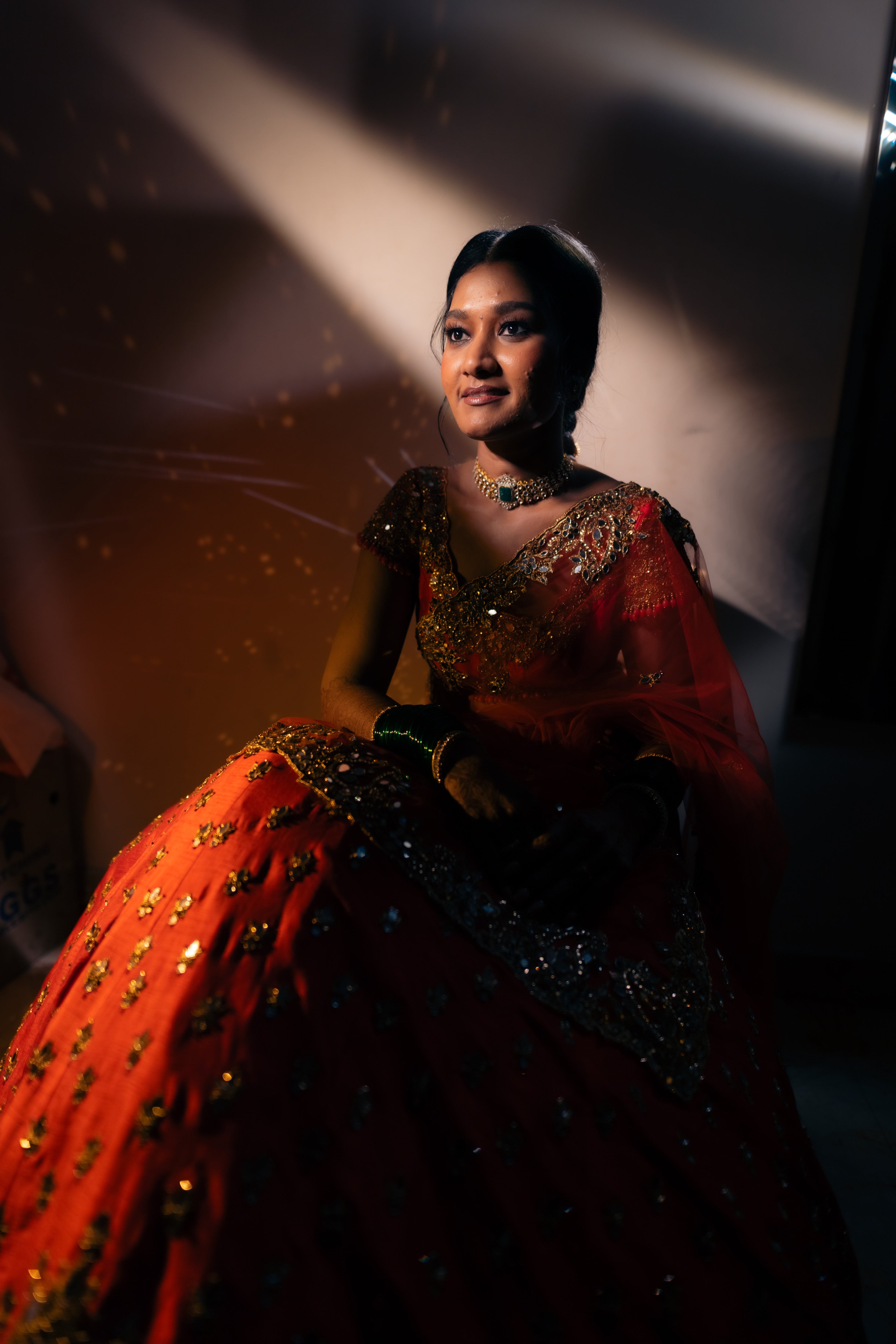 Bride Vaishali from Dubai, in a stunning mirror and zardozi worked orange lehenga designed by Archana Karthick. Here the bride is posing in a seated position looking slightly away from the camera.