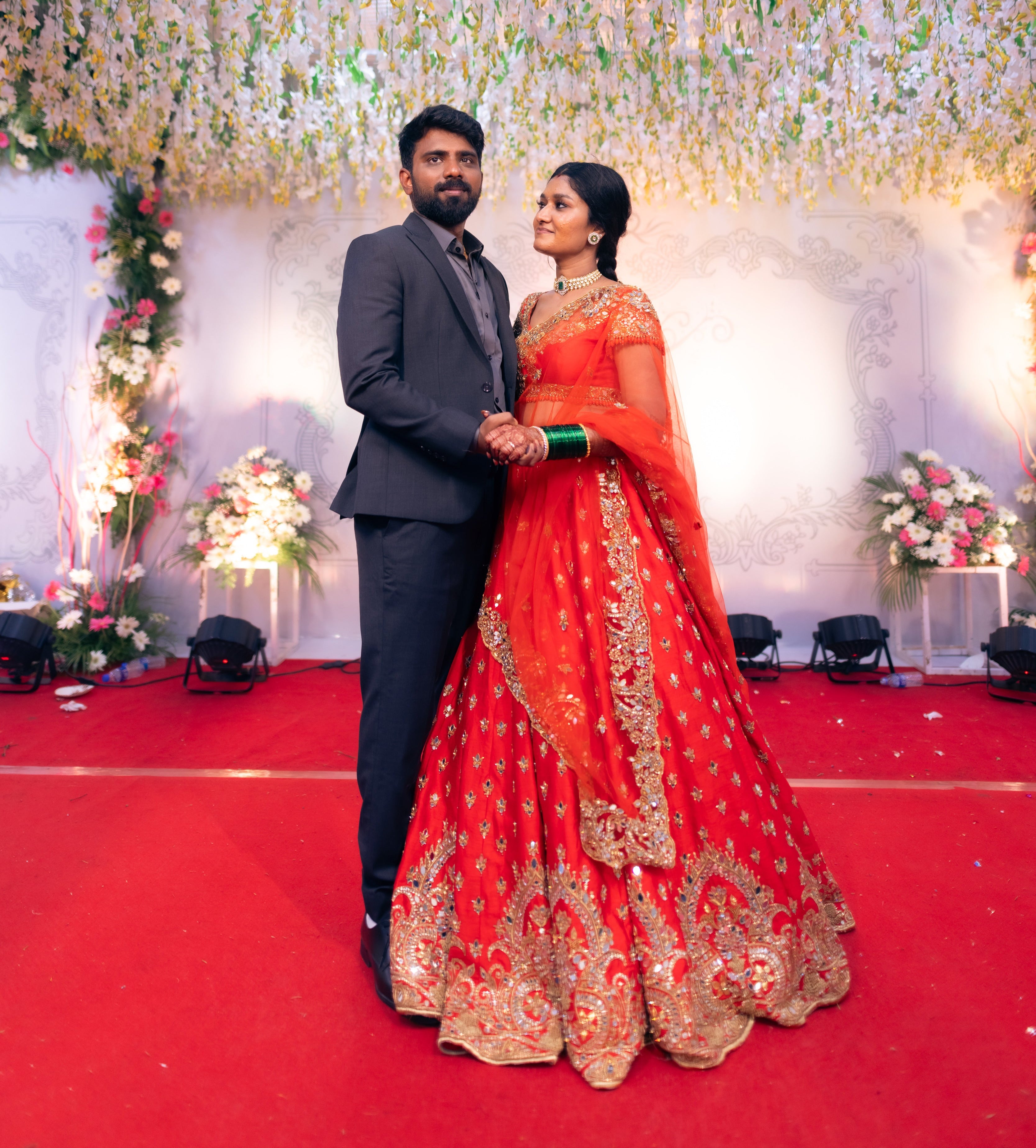 Bride Vaishali from Dubai, in a stunning mirror and zardozi worked orange lehenga designed by Archana Karthick. Here the bride is posing with the groom on the stage looking at the groom.