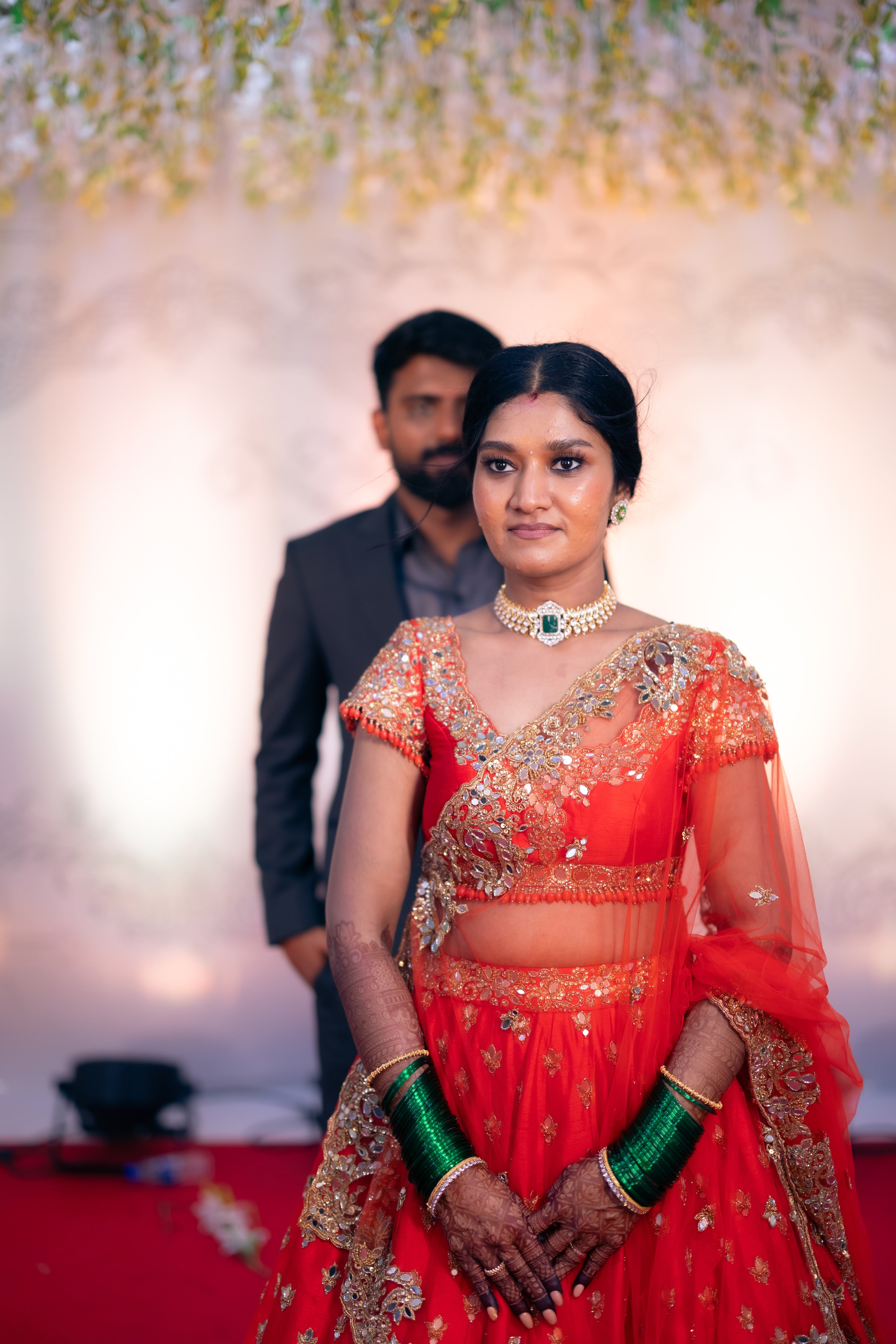 Bride Vaishali from Dubai, in a stunning mirror and zardozi worked orange lehenga designed by Archana Karthick. Here the bride is in front of the groom on the stage posing for a half front profile.
