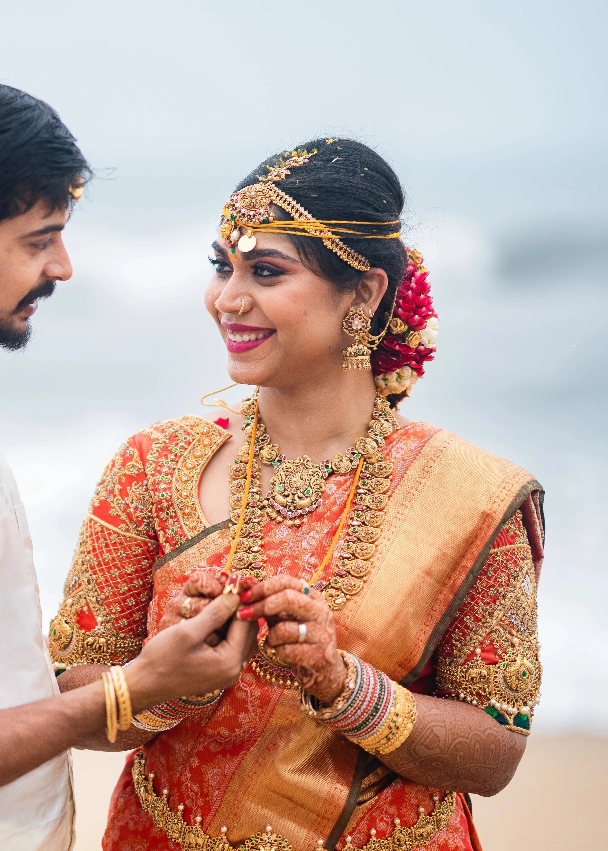 Bride Vidya D smiling with groom in an orange silk blouse, handcrafted with beads, zardozi, kundan, resham, stones and zardozi by team Archana Karthick.