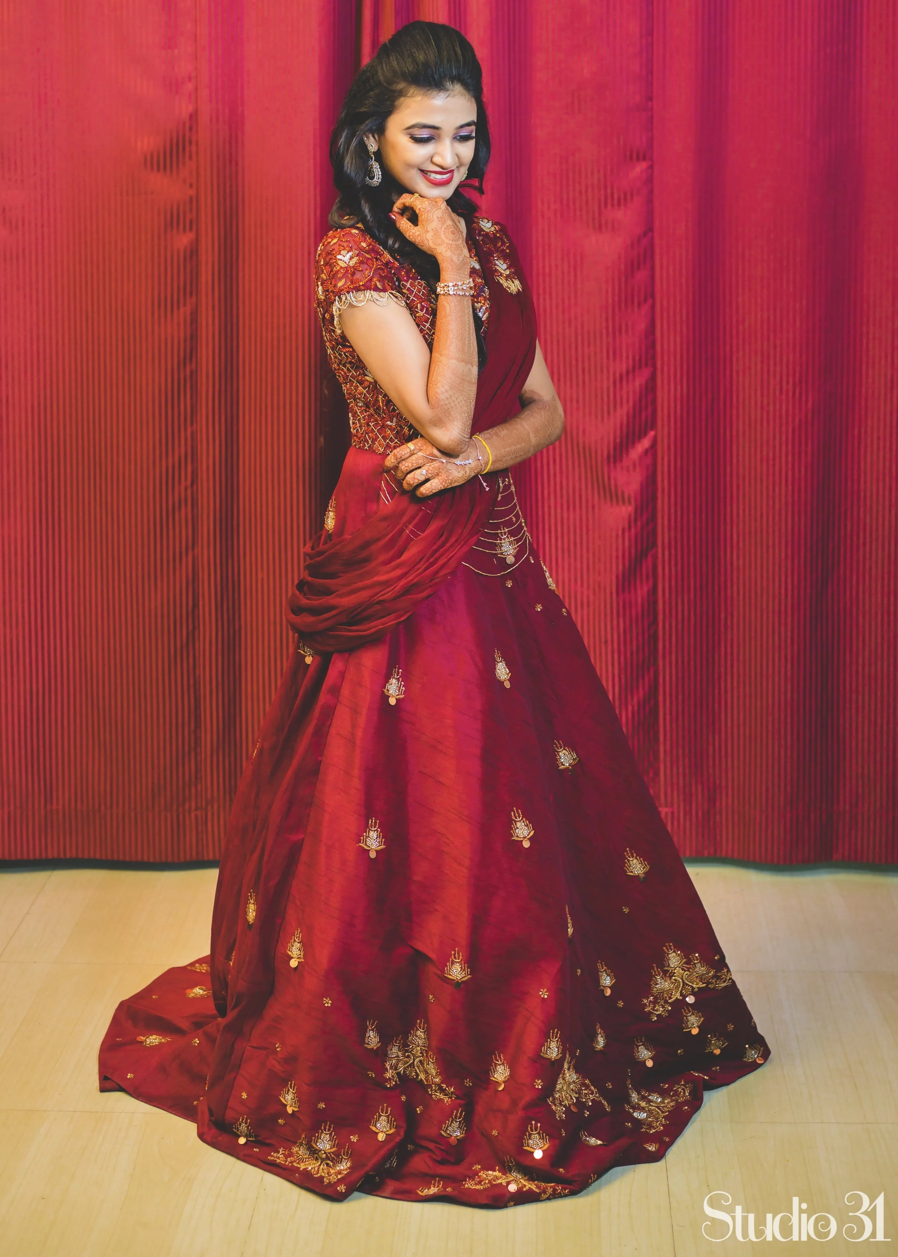 Bride from Chennai looking down and smiling in a handcrafted maroon raw silk gown with draped dupatta.