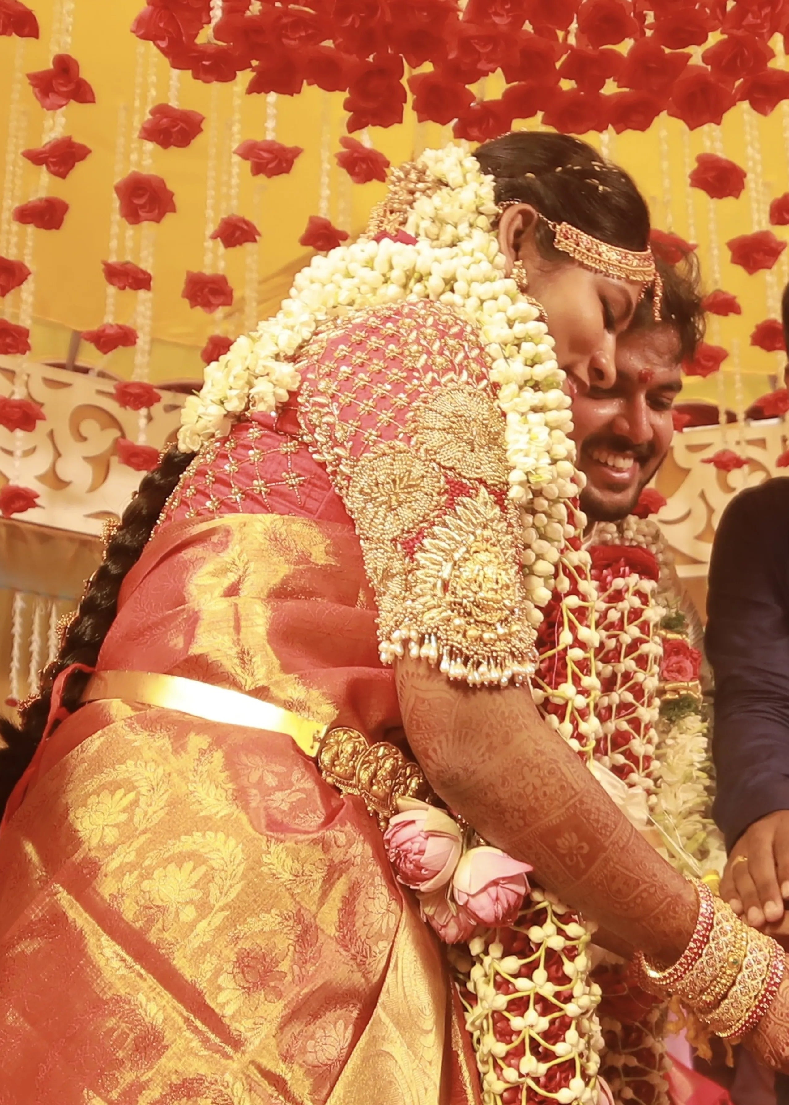 Bride from Chennai posing with groom in a handcrafted red raw silk blouse embellished with zardozi, pearls, beads with goddess pendants on sleeves.