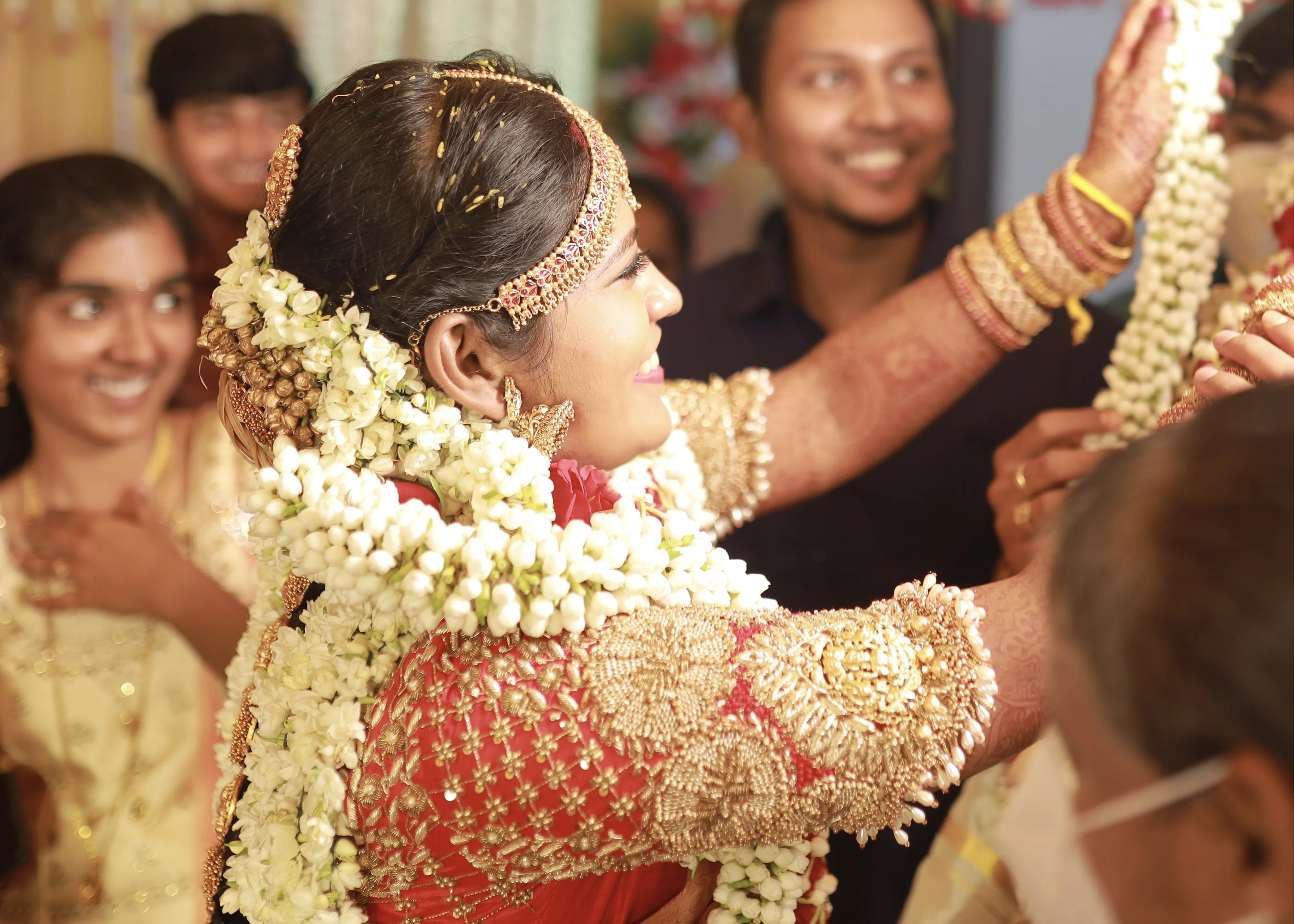 Bride from Chennai smiling in a handcrafted red raw silk blouse embellished with zardozi, pearls, beads with goddess pendants on sleeves.