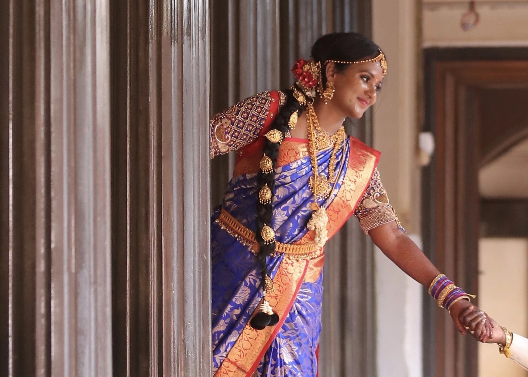 Bride posing for photoshoot in a concept peacock blouse in blue and red silk, handcrafted with zardozi, beads, resham knot work and goddess pendant on sleeves.