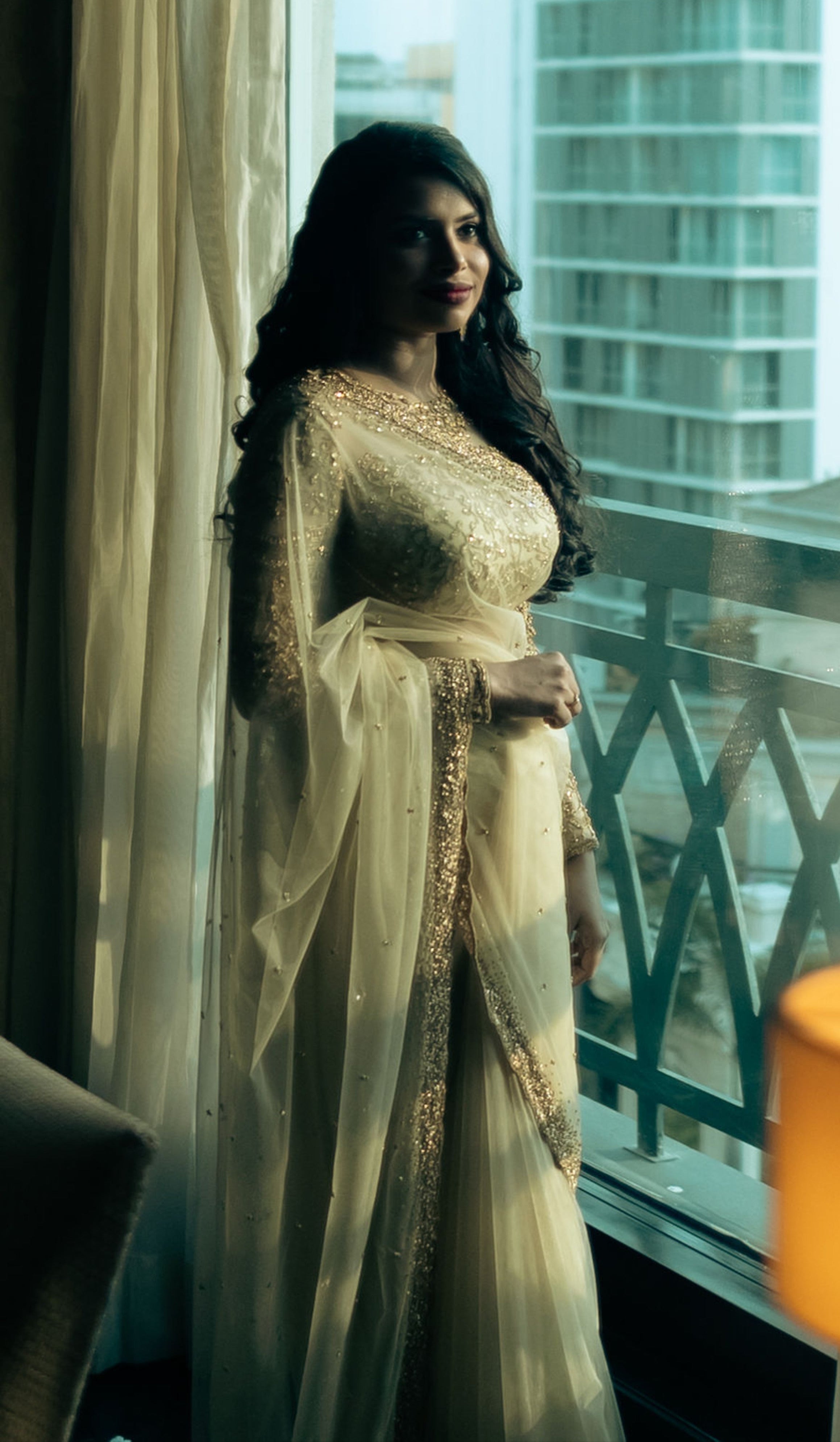 Christian Bride Steffi in handcrafted net saree in champagne gold designed by Archana Karthick. Posing by the window for a full side profile view.