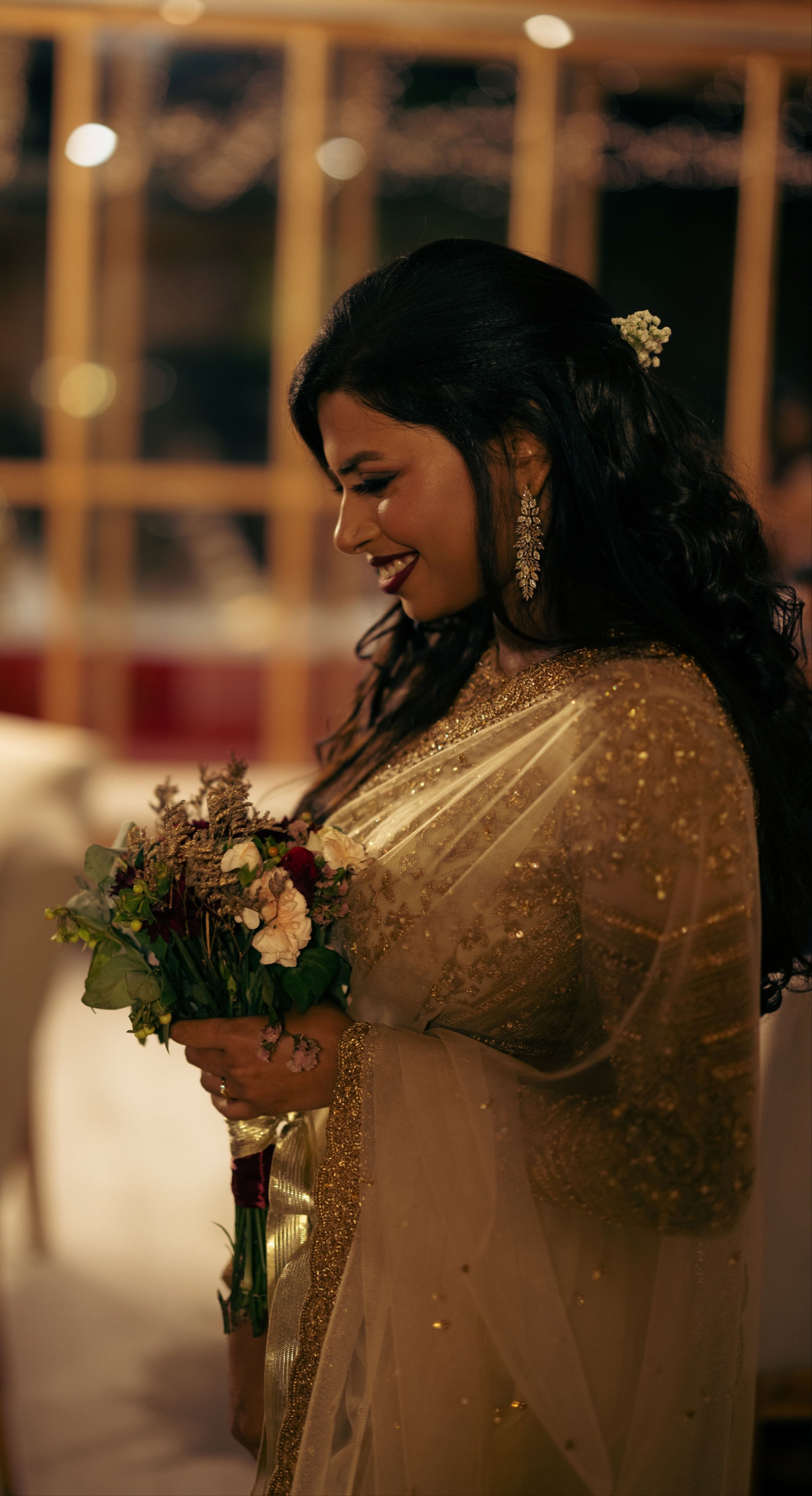Christian Bride Steffi in handcrafted net saree in champagne gold designed by Archana Karthick. Posing in a shy pose looking down with a bouquet in hand. half side profile.
