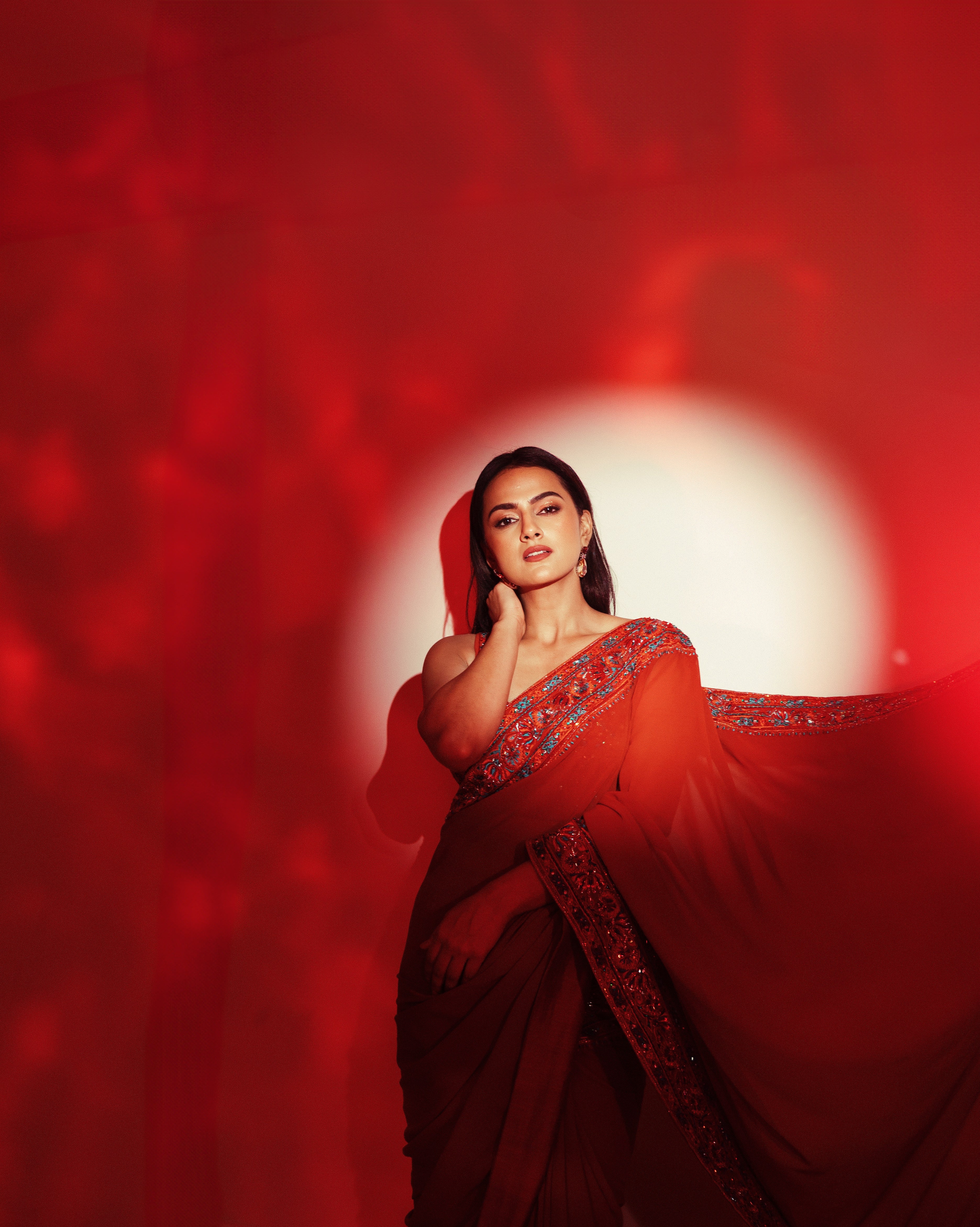 Indian film actress Shraddha Srinath poses in a burnt orange saree by Archana Karthick, featuring intricate embroidery on the blouse and borders, paired with elegant gold earrings against a bright orange backdrop.