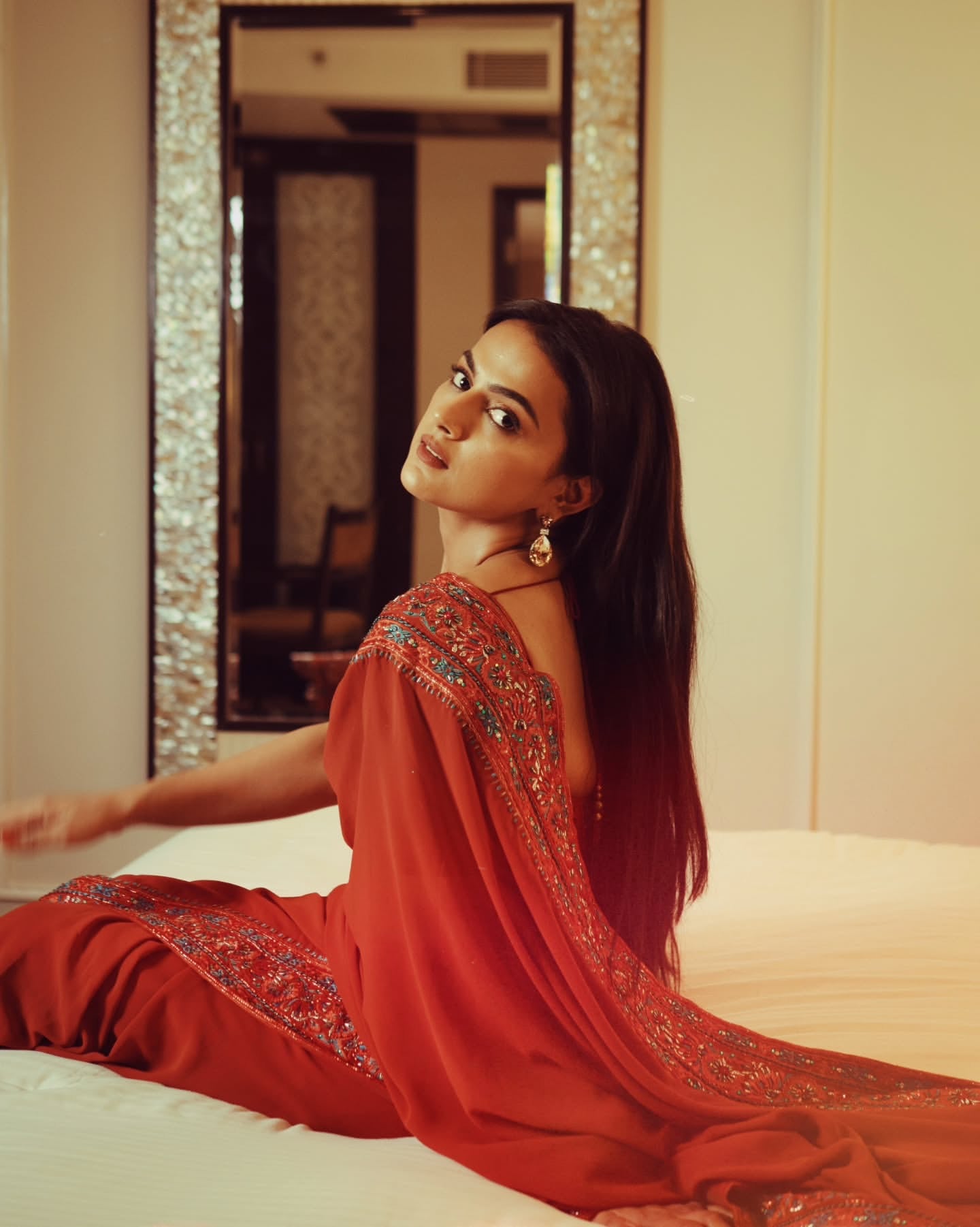 Indian film actress Shraddha Srinath poses seated on the bed, in a burnt orange saree by Archana Karthick, featuring intricate embroidery on the blouse and borders, against the backdrop of a large mirror.
