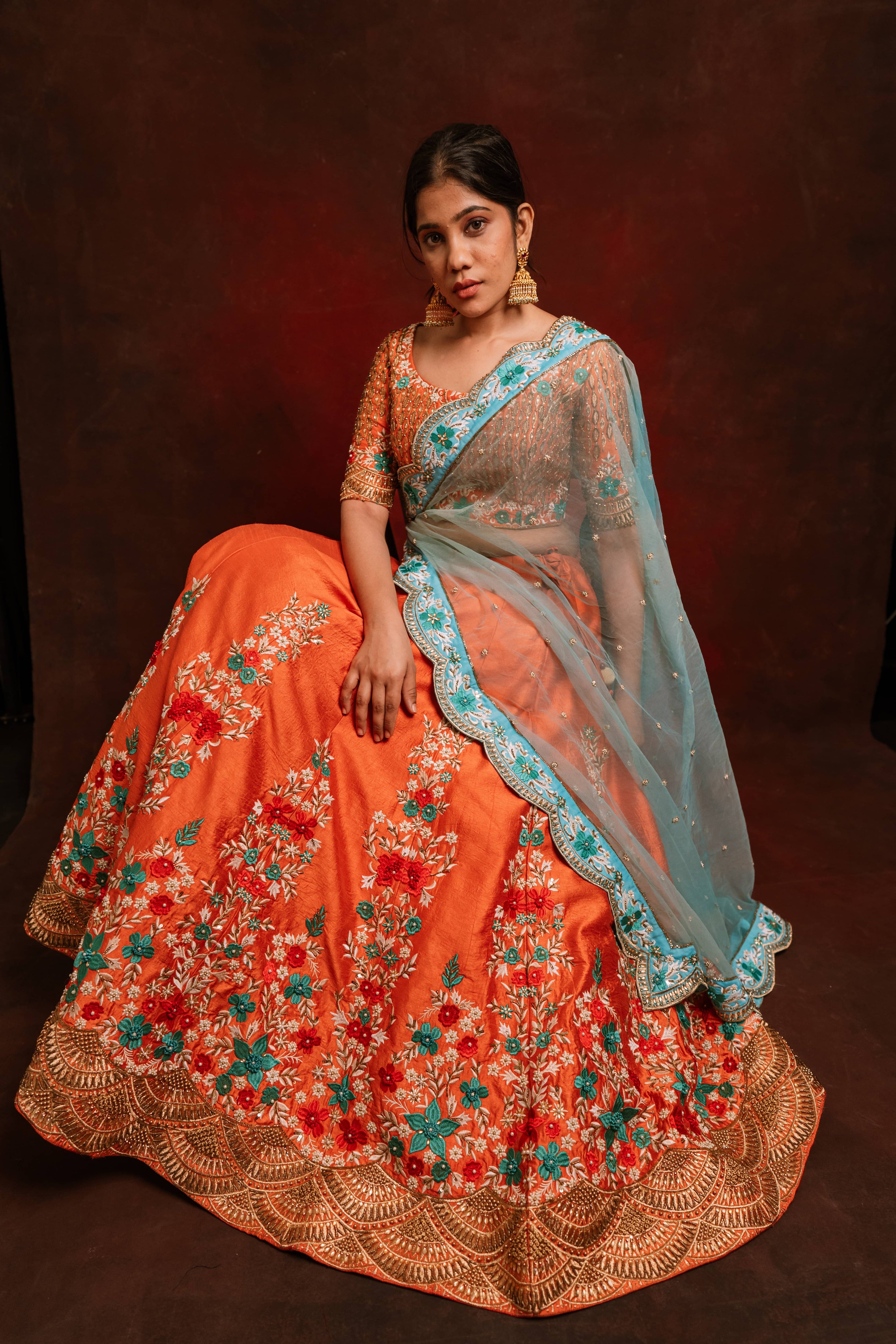 Jaina Parmar, known for recreating looks of actress Nayanthara is seen here posing in a raw silk burnt orange lehenga with blue net dupatta. seated pose looking at camera