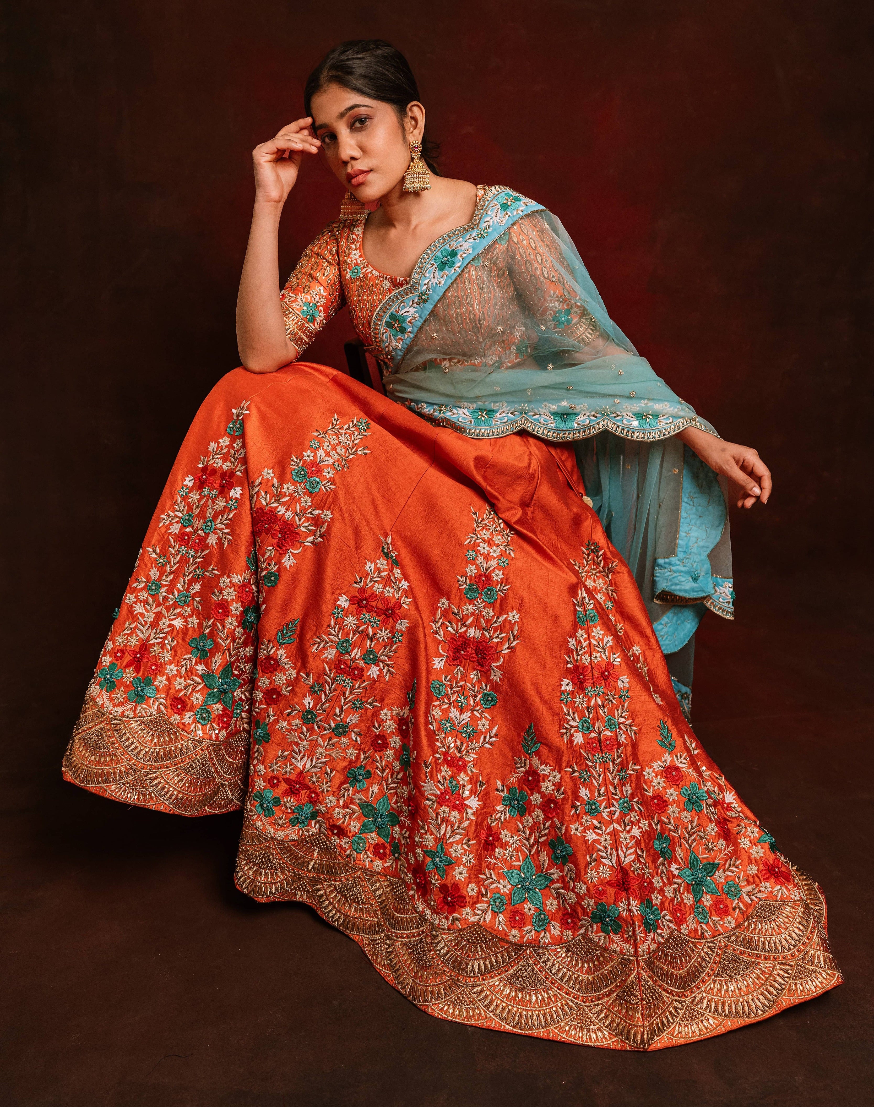 Jaina Parmar, known for recreating looks of actress Nayanthara is seen here posing in a raw silk burnt orange lehenga with blue net dupatta. seated pose with hand on forehead.