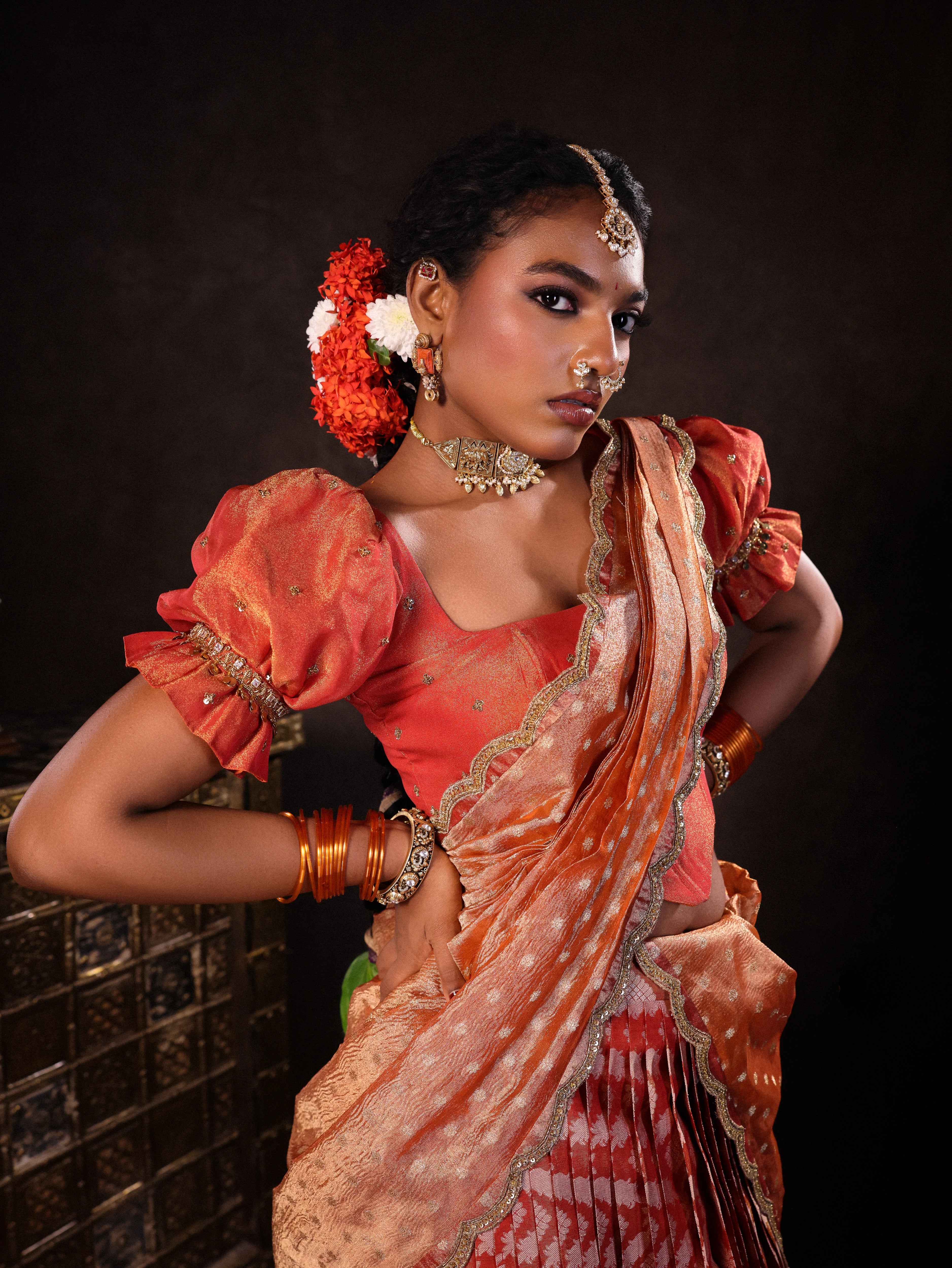 Model Ashika Crystal posing during photoshoot in a puff sleeved orange half saree set, handcrafted with zardozi, sequins, metal coin hangings and square cut stones. Team Archana Karthick completed the style with orange glass bangles, a ring, jhumkas, nose ring and a maang tikka.