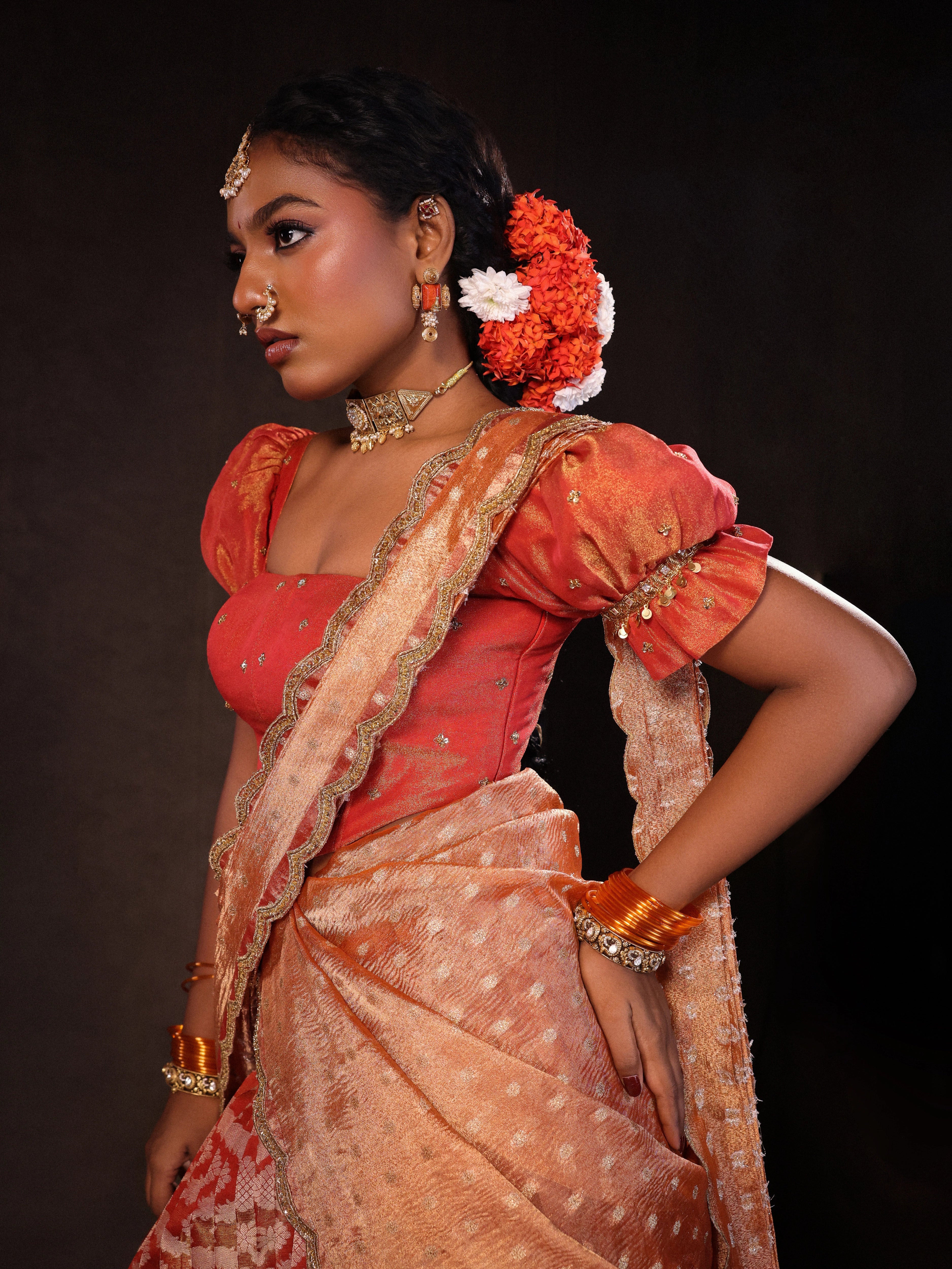 Model Ashika Crystal posing for a side profile in a puff sleeved orange half saree set, handcrafted with zardozi, sequins, metal coin hangings and square cut stones. Team Archana Karthick completed the style with orange glass bangles, a ring, jhumkas, nose ring and a maang tikka.