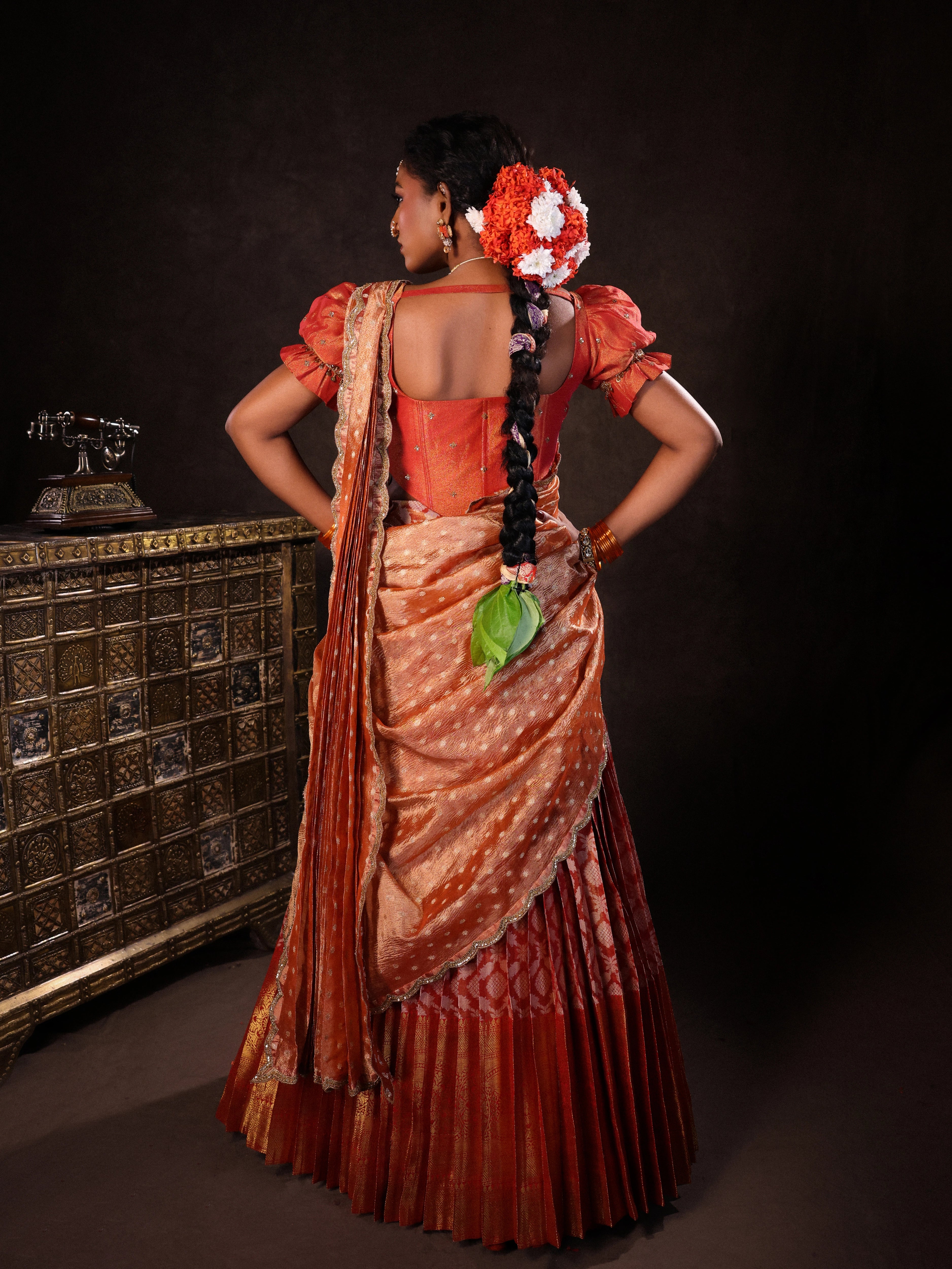 Model Ashika Crystal posing for back design in a puff sleeved orange half saree set, handcrafted with zardozi, sequins, metal coin hangings and square cut stones. Team Archana Karthick completed the style with orange glass bangles, a ring, jhumkas, nose ring and a maang tikka.