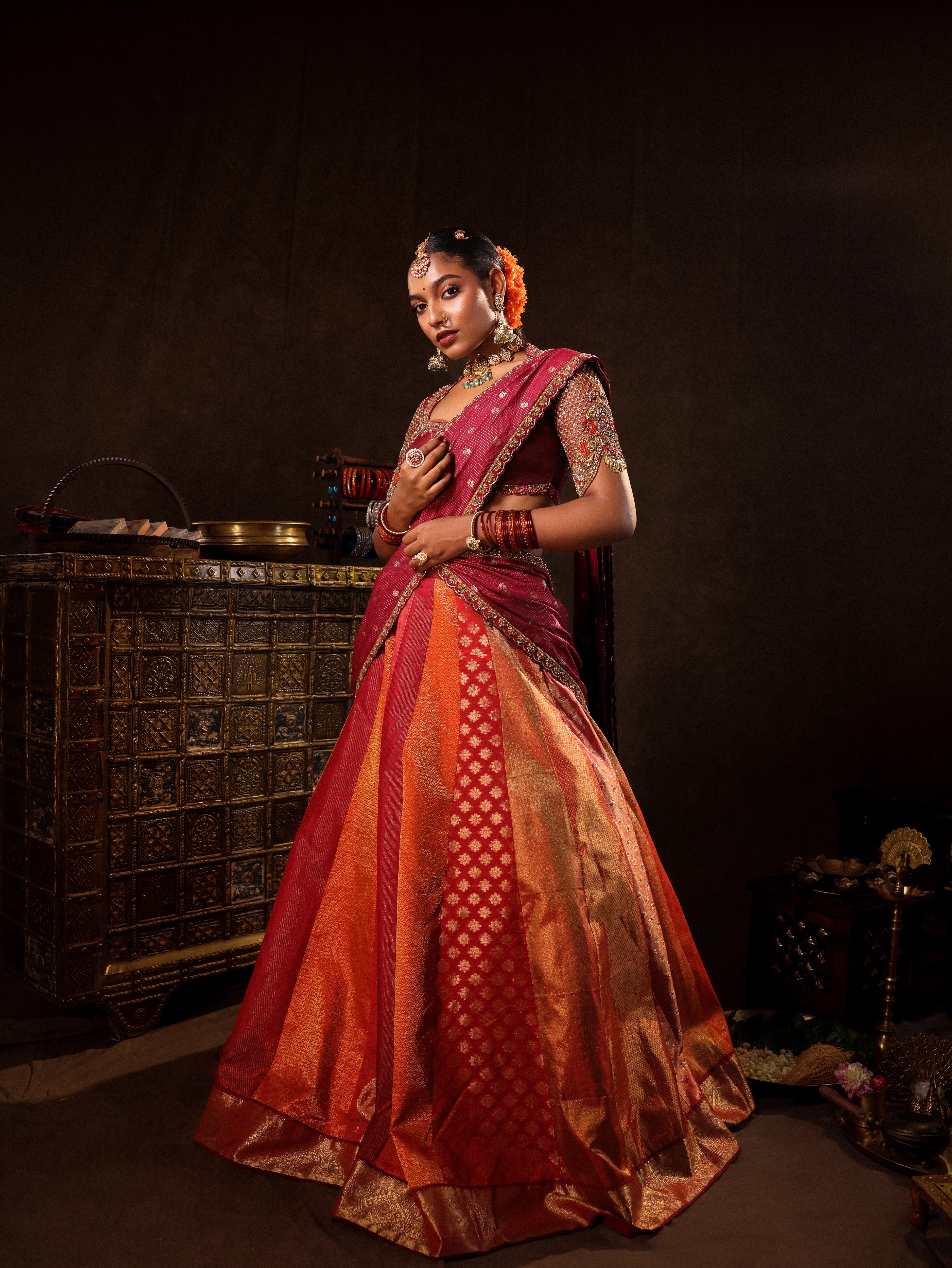 Model Ashika Crystal posing for photoshoot in a red and orange silk half saree with a scalloped veil is handrcrafted with sequins, cutdana, resham, stones and exquisite hangings. Team Archana Karthick completed the style with orange & red glass bangles, a ring, jhumkas, nose ring and a maang tikka.