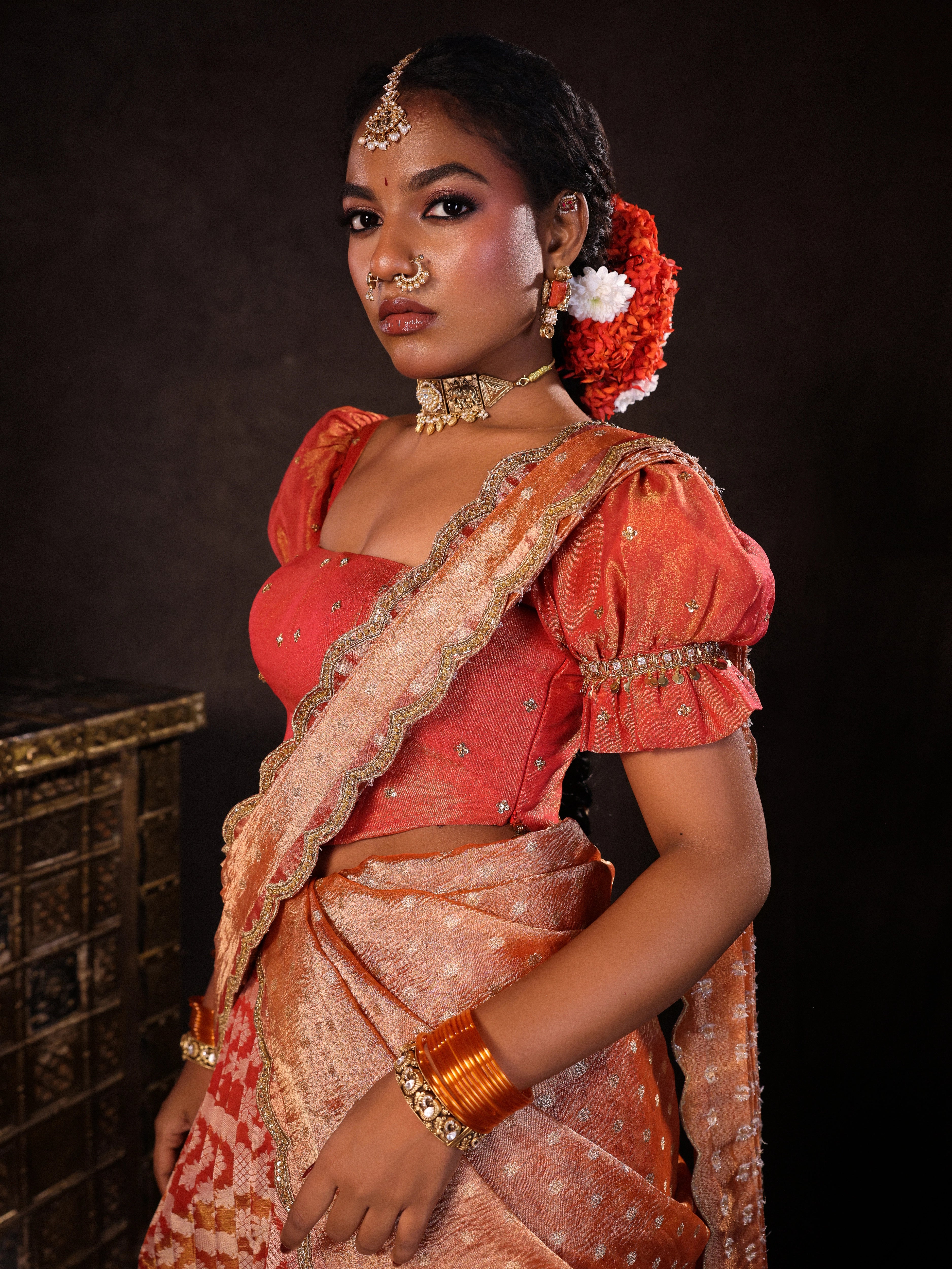 Model Ashika Crystal posing for side profile in a puff sleeved orange half saree set, handcrafted with zardozi, sequins, metal coin hangings and square cut stones. Team Archana Karthick completed the style with orange glass bangles, a ring, jhumkas, nose ring and a maang tikka.