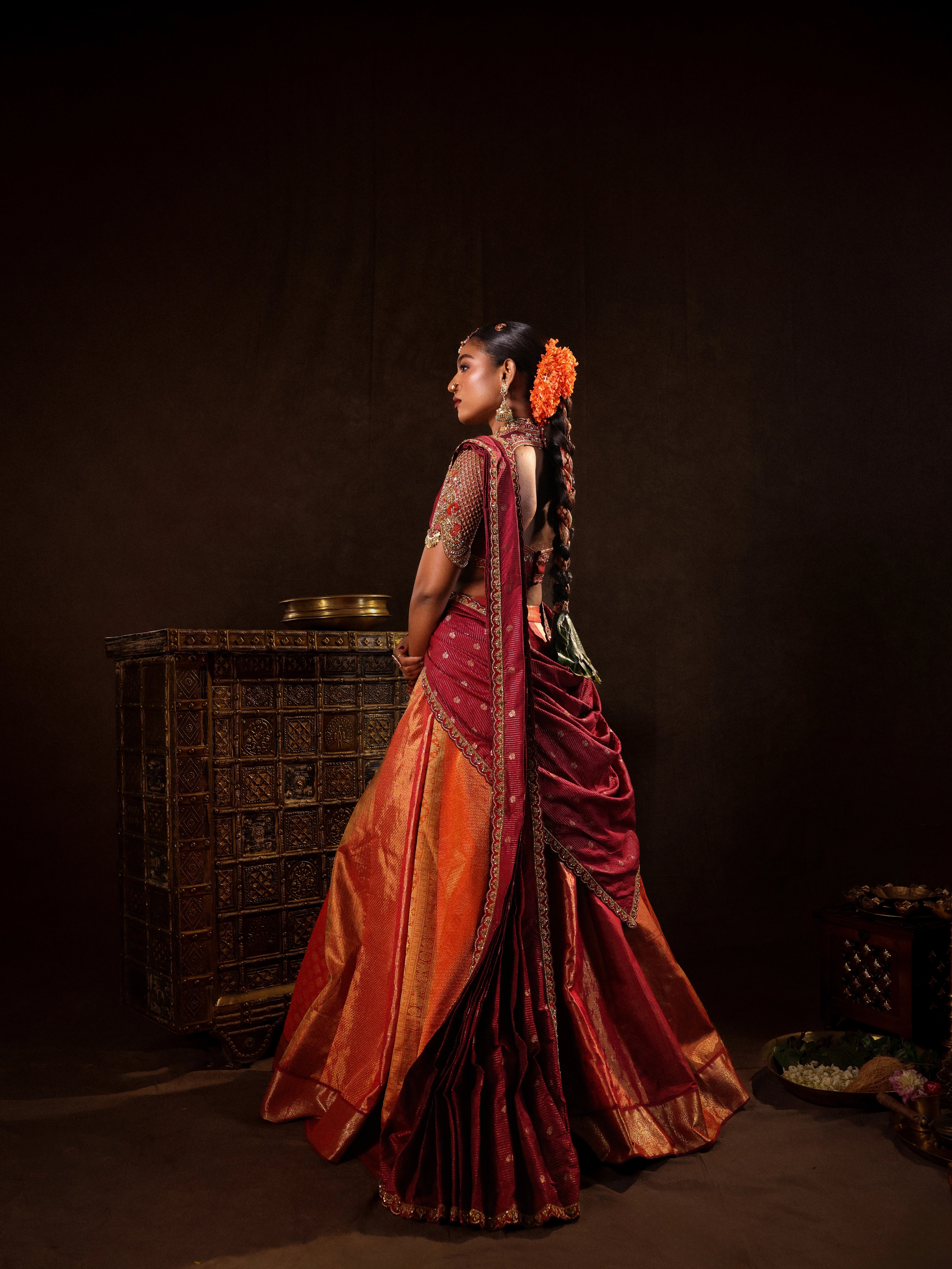 Model Ashika Crystal posing for side profile view in a red and orange silk half saree with a scalloped veil is handrcrafted with sequins, cutdana, resham, stones and exquisite hangings. Team Archana Karthick completed the style with orange & red glass bangles, a ring, jhumkas, nose ring and a maang tikka.
