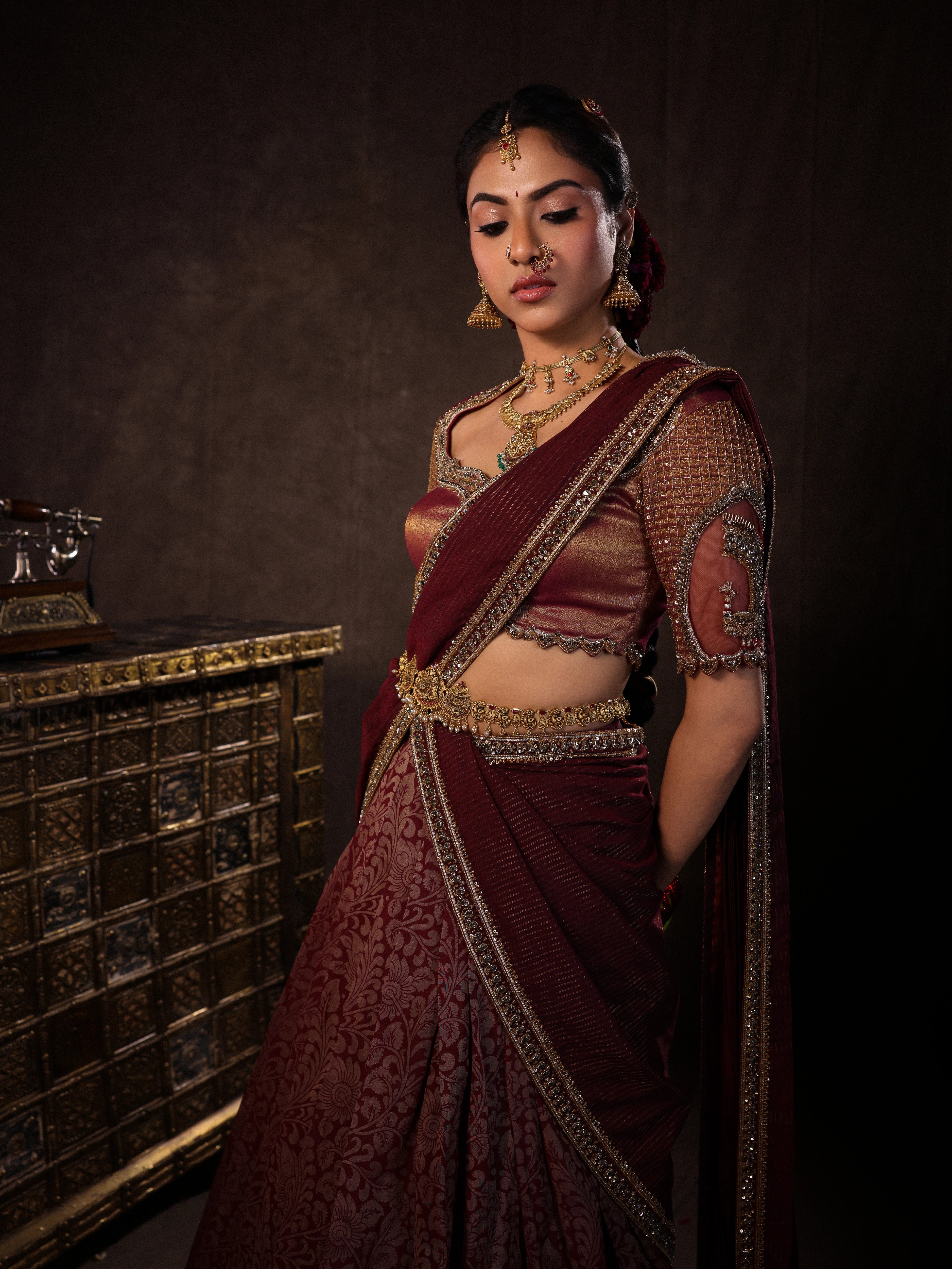 Model Dr Sree Advaita posing during photoshoot in a brownish maroon silk half saree set intricately handcrafted with zardozi, resham, cutdana, sequins & rhinestones. Team Archana Karthick completed the style with maroon glass bangles, a ring, jhumkas, nose ring and a maang tikka.