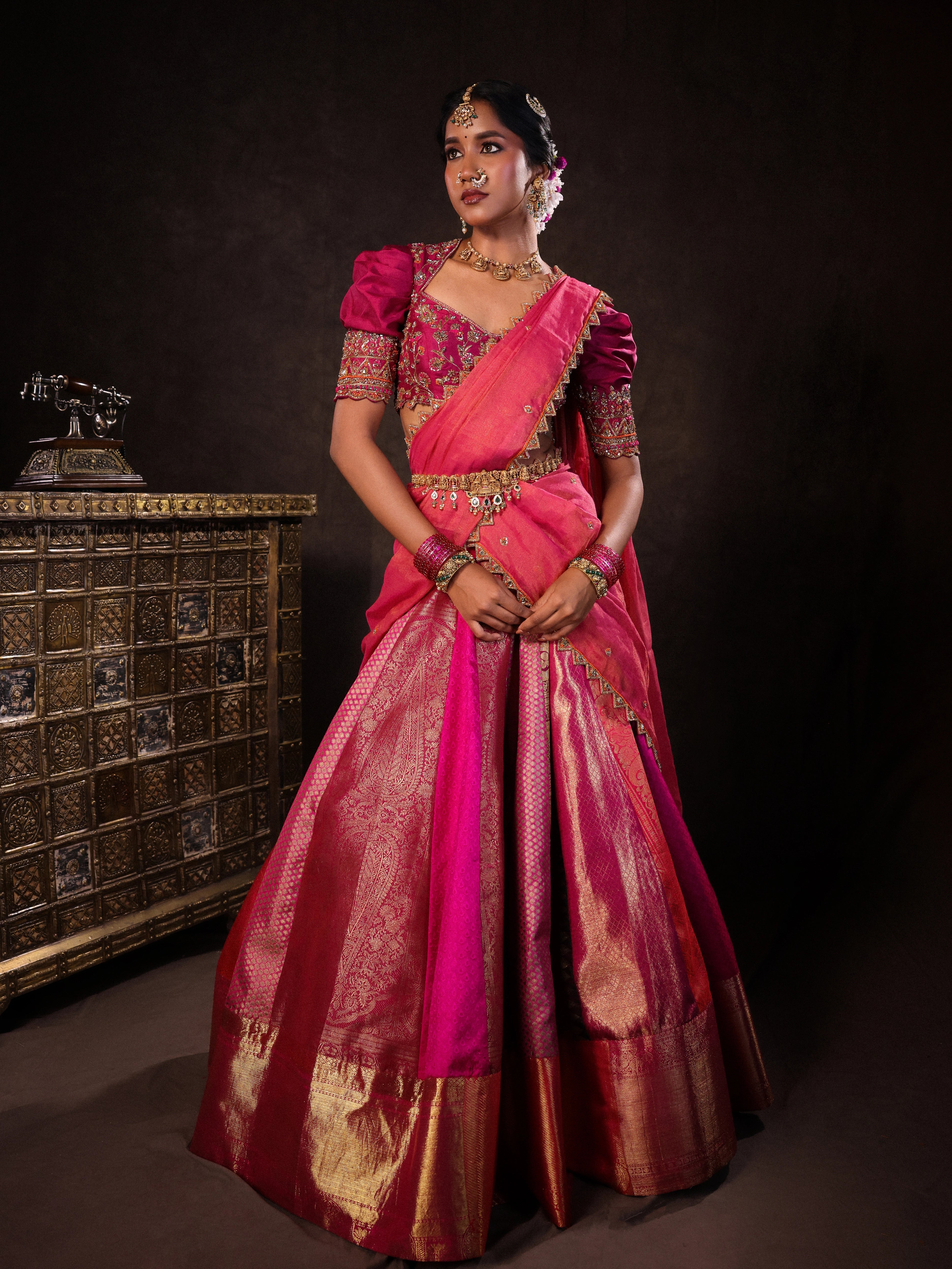 Model Gayathri Dhanasekar in a pink & orange silk half saree handcrafted with zardozi, cutdana (bugle beads), resham (silk thread), sequins and rhinestones. Team Archana Karthick completed the style with pink glass bangles, a ring, a choker, jhumkas, nose ring and a maang tikka.