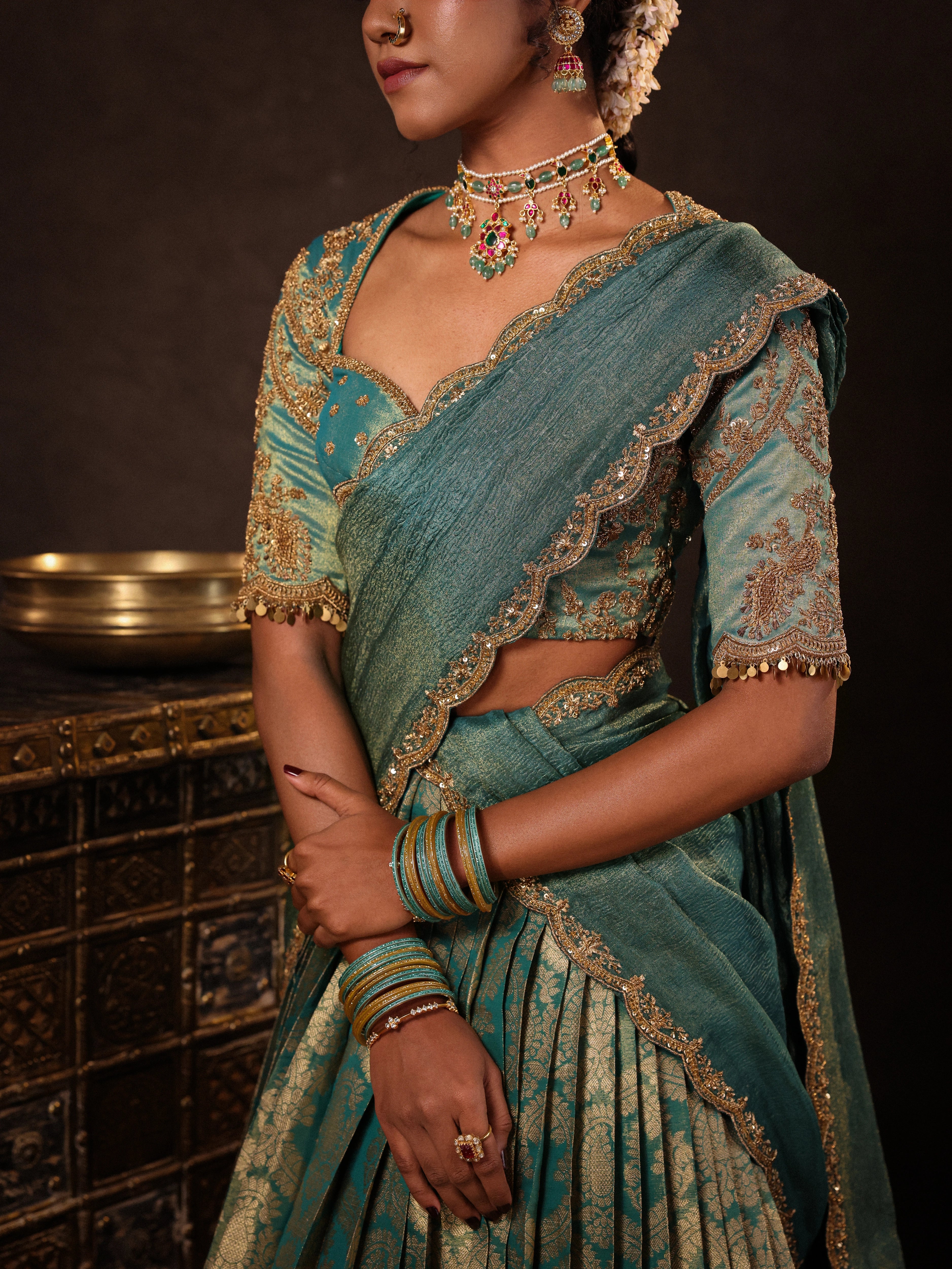 Model Gayathri Dhanasekar posing during photoshoot in a teal half saree (silk blouse and skirt and tissue dupatta) handcrafted with zardozi, zari, cutdana, sequins and metal coin hangings. Team Archana Karthick completed the style with teal glass bangles, a ring, a choker, jhumkas, nose ring and a maang tikka.