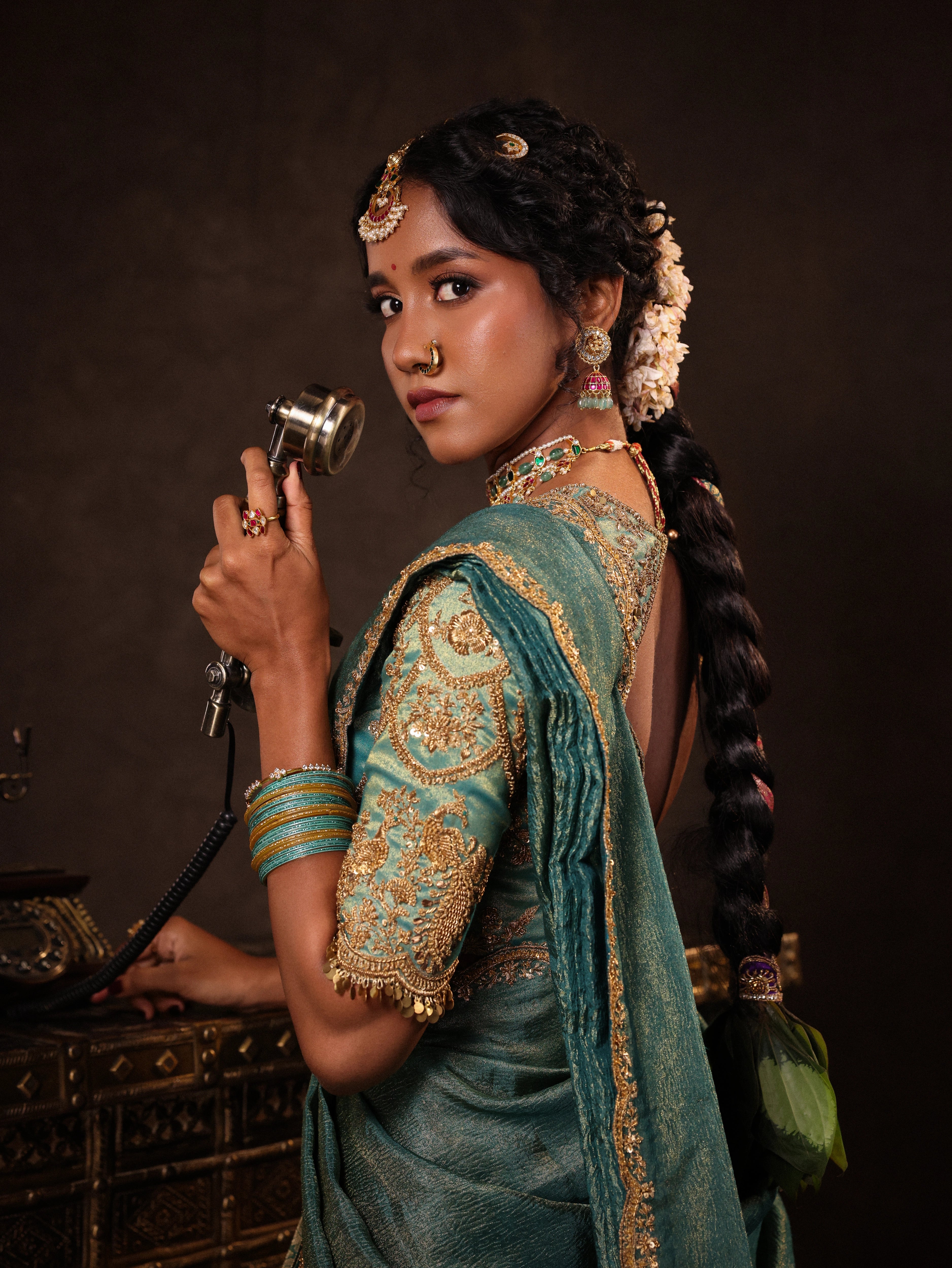 Model Gayathri Dhanasekar posing during photoshoot with a vintage phone in a teal half saree (silk blouse and skirt and tissue dupatta) handcrafted with zardozi, zari, cutdana, sequins and metal coin hangings. Team Archana Karthick completed the style with teal glass bangles, a ring, a choker, jhumkas, nose ring and a maang tikka.