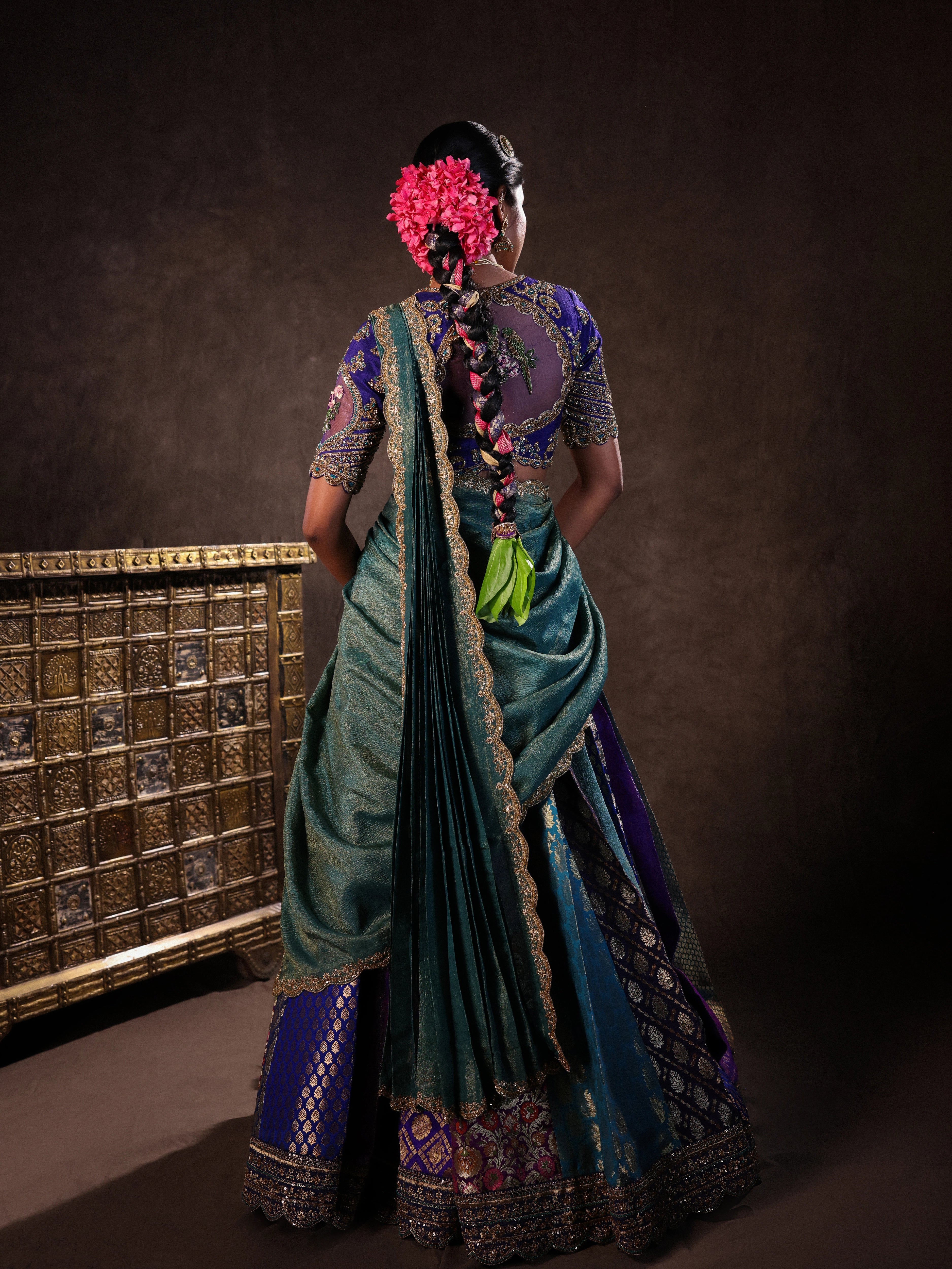 Model Gayathri Dhanasekar posing for back design in a brocade, raw silk and tissue silk half saree set in shades of blue handcrafted with zardozi, resham, sequins and rhinestones. Team Archana Karthick completed the style with blue glass bangles, a ring, a choker, jhumkas, nose ring and a maang tikka.
