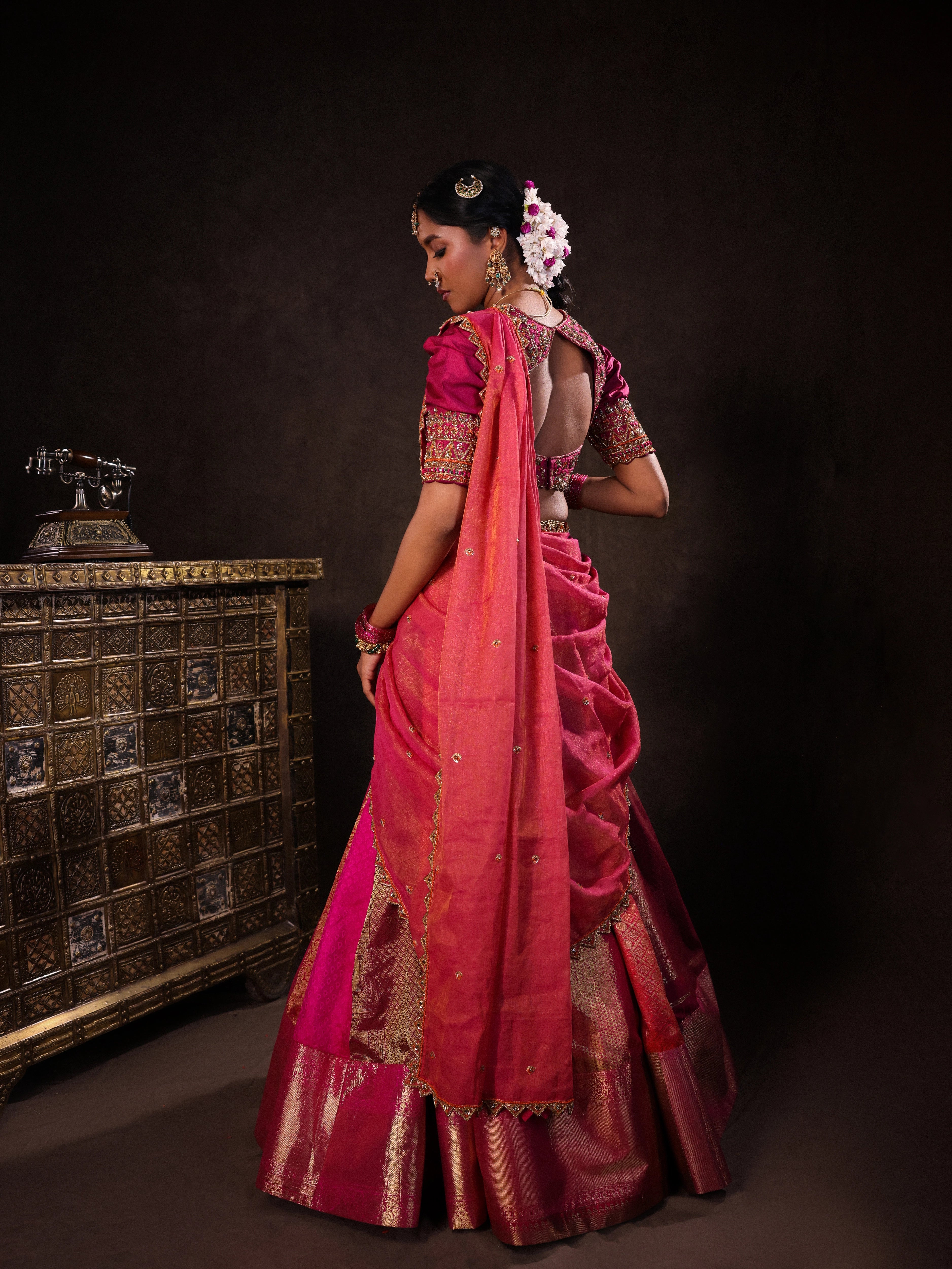 Model Gayathri Dhanasekar posing for back design in a pink & orange silk half saree handcrafted with zardozi, cutdana (bugle beads), resham (silk thread), sequins and rhinestones. Team Archana Karthick completed the style with pink glass bangles, a ring, a choker, jhumkas, nose ring and a maang tikka.