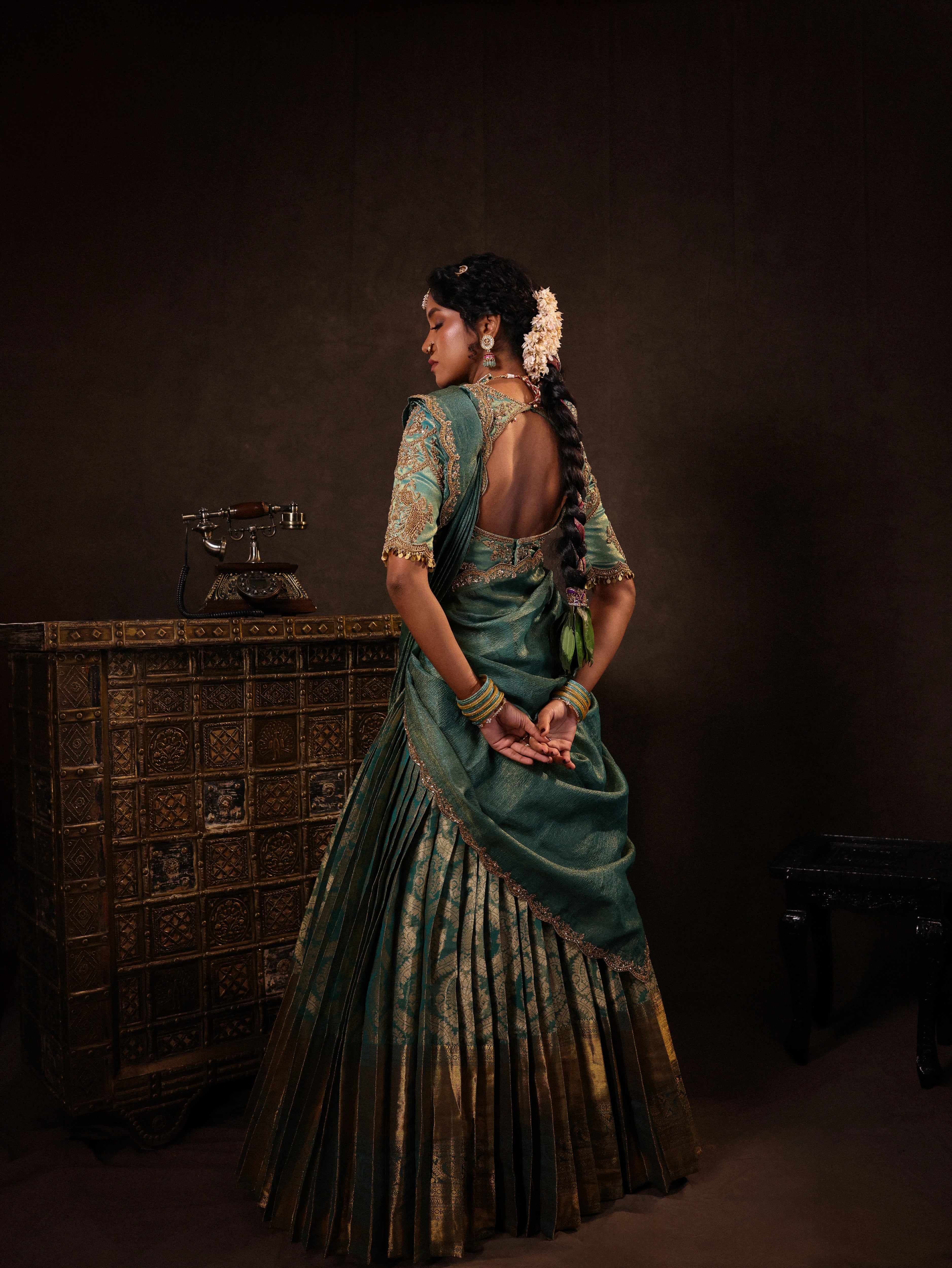 Model Gayathri Dhanasekar posing for back design in a teal half saree (silk blouse and skirt and tissue dupatta) handcrafted with zardozi, zari, cutdana, sequins and metal coin hangings. Team Archana Karthick completed the style with teal glass bangles, a ring, a choker, jhumkas, nose ring and a maang tikka.