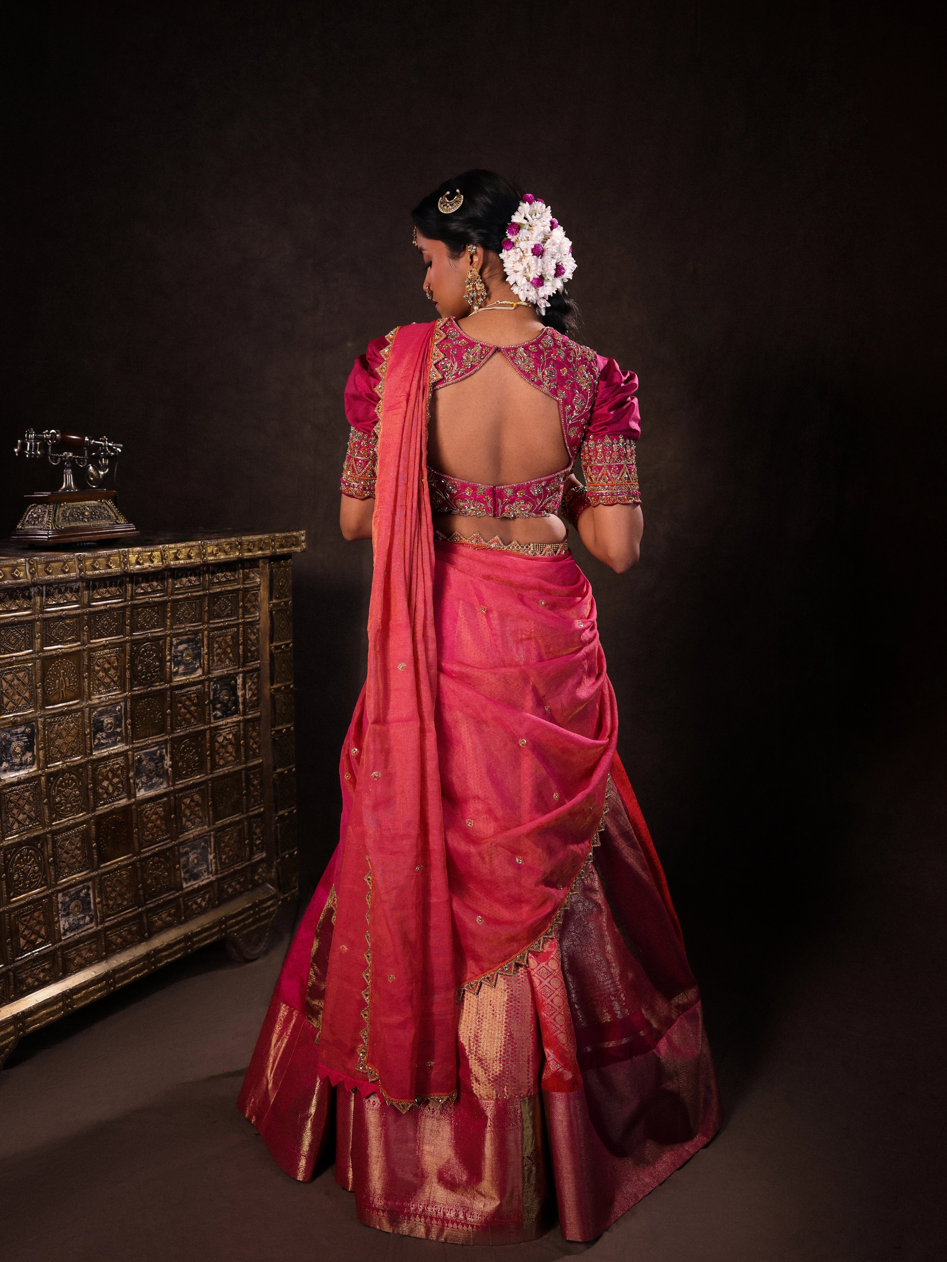 Model Gayathri Dhanasekar posing for back detailing in a pink & orange silk half saree handcrafted with zardozi, cutdana (bugle beads), resham (silk thread), sequins and rhinestones. Team Archana Karthick completed the style with pink glass bangles, a ring, a choker, jhumkas, nose ring and a maang tikka.