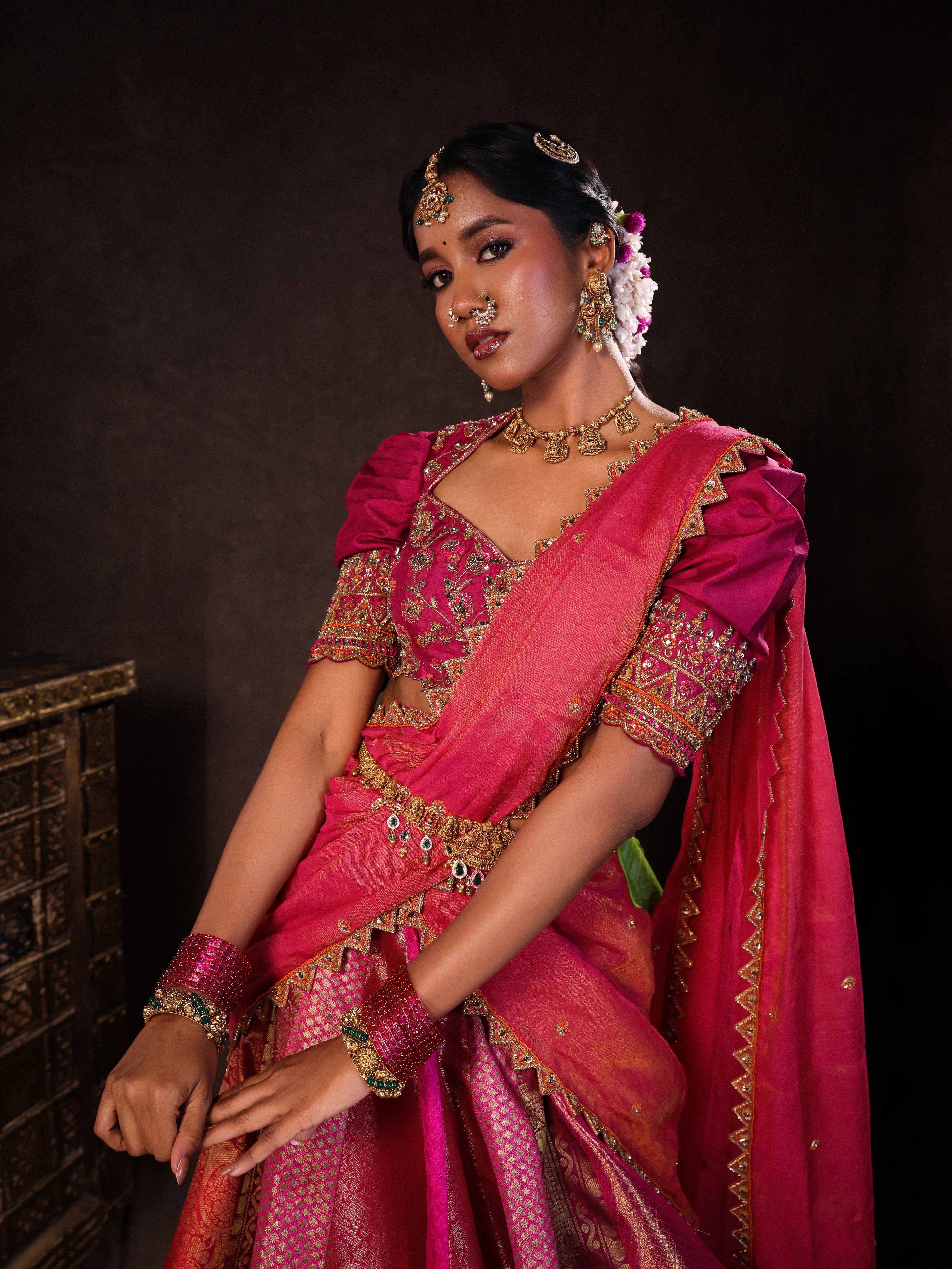 Model Gayathri Dhanasekar posing in a photoshoot in a pink & orange silk half saree handcrafted with zardozi, cutdana (bugle beads), resham (silk thread), sequins and rhinestones. Team Archana Karthick completed the style with pink glass bangles, a ring, a choker, jhumkas, nose ring and a maang tikka.